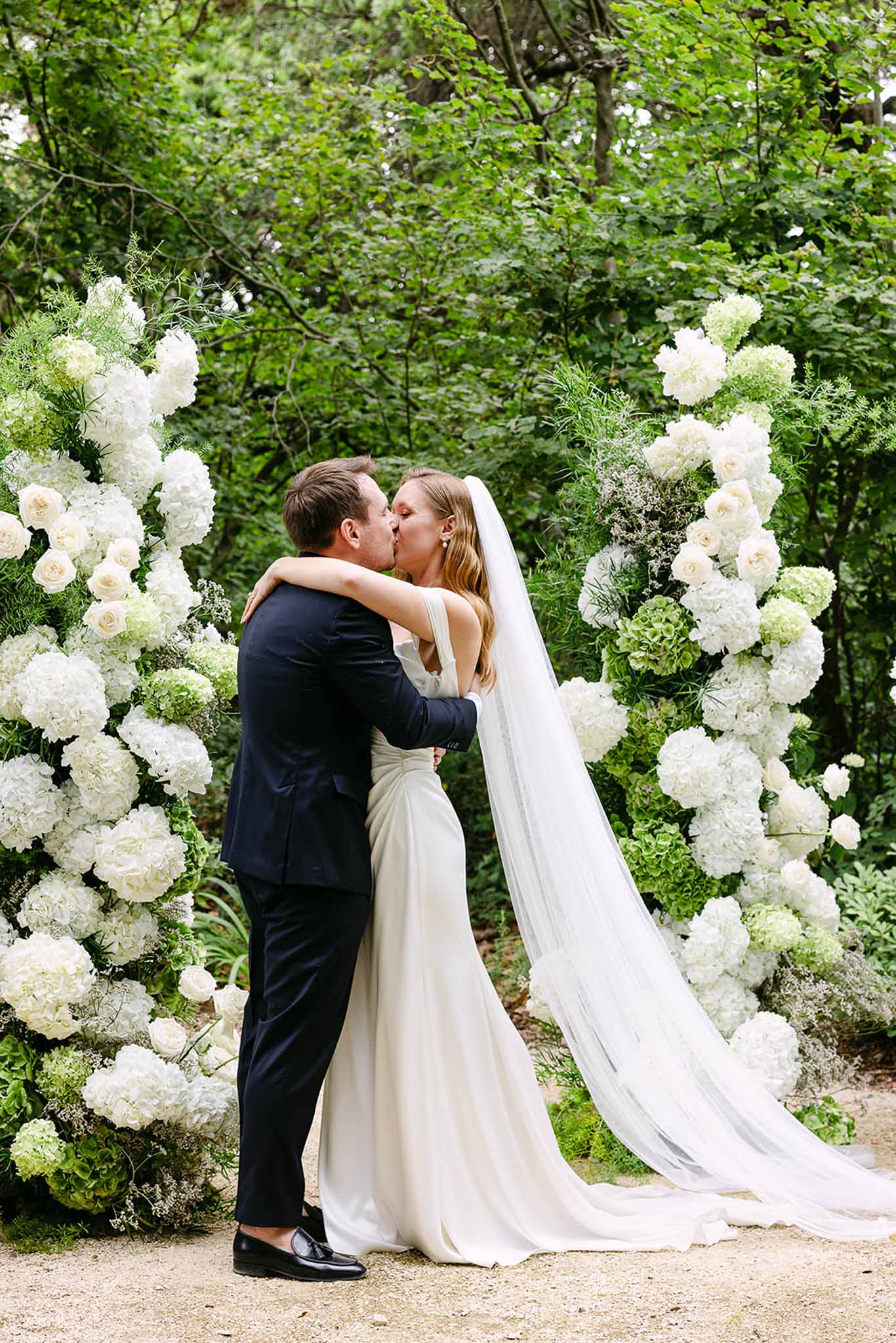 First kiss under white hydrangea and ivory rose column arch with bride's cathedral veil trailing behind