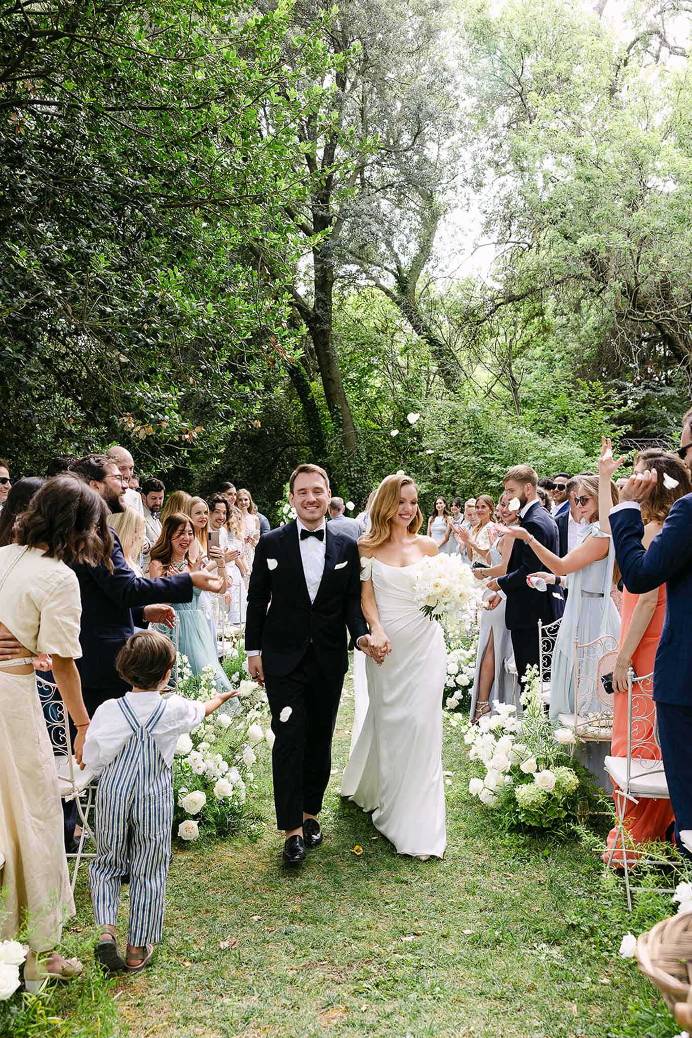 Newlyweds walk down garden aisle as guests toss white petals with white floral arrangements lining path