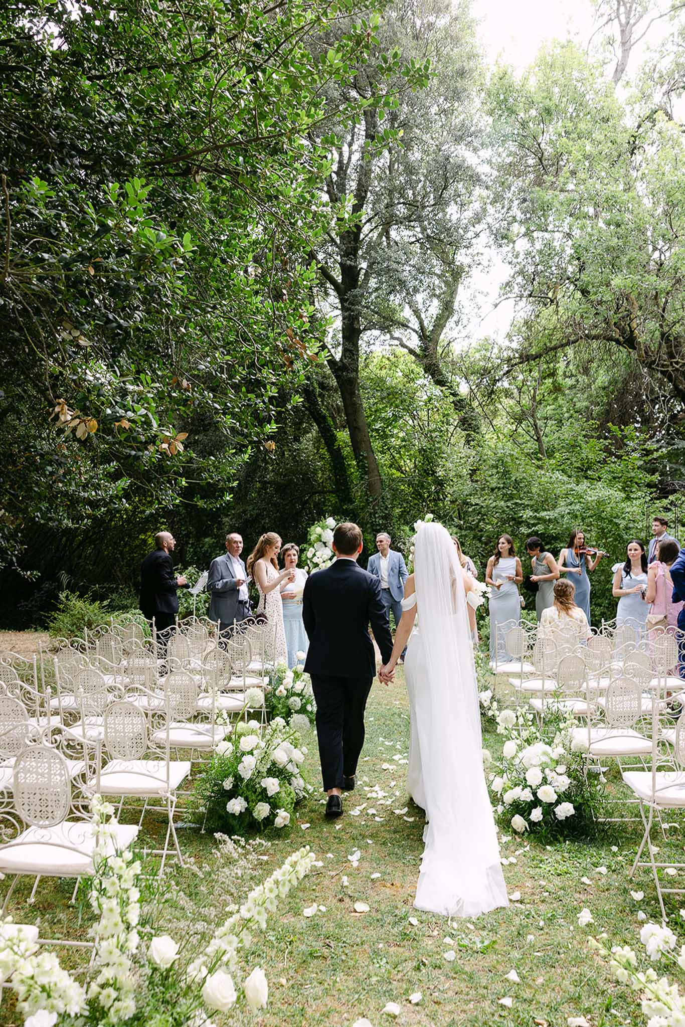 Couple walks back down petal-strewn grass aisle past white rose arrangements with bridesmaids in dusty blue