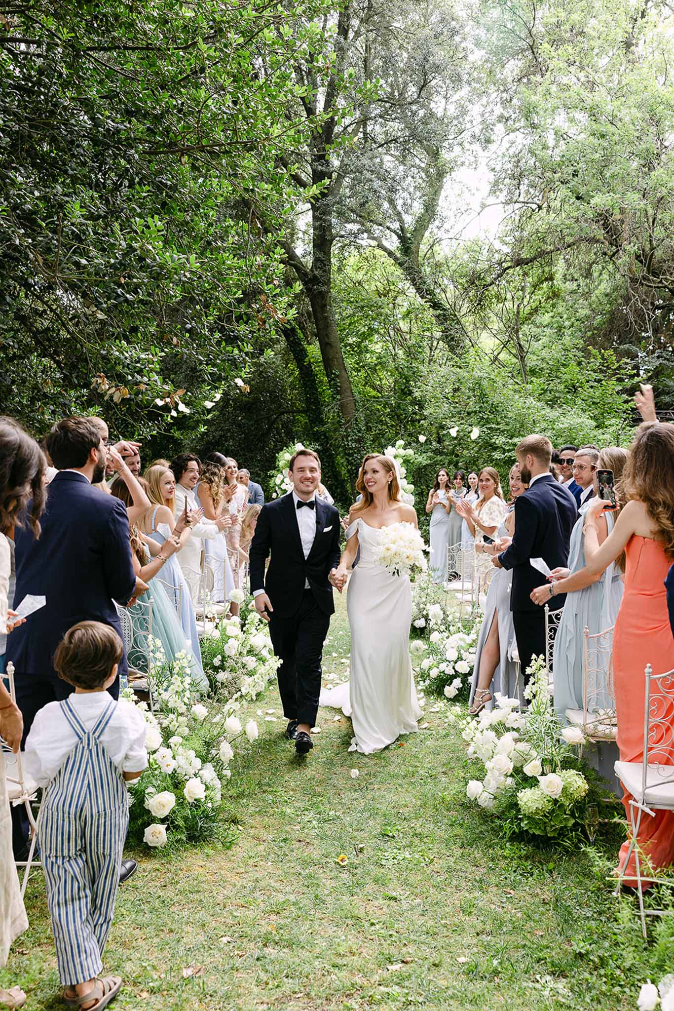 Bride and groom walk aisle as guests toss white petals past peony ground arrangements on garden lawn
