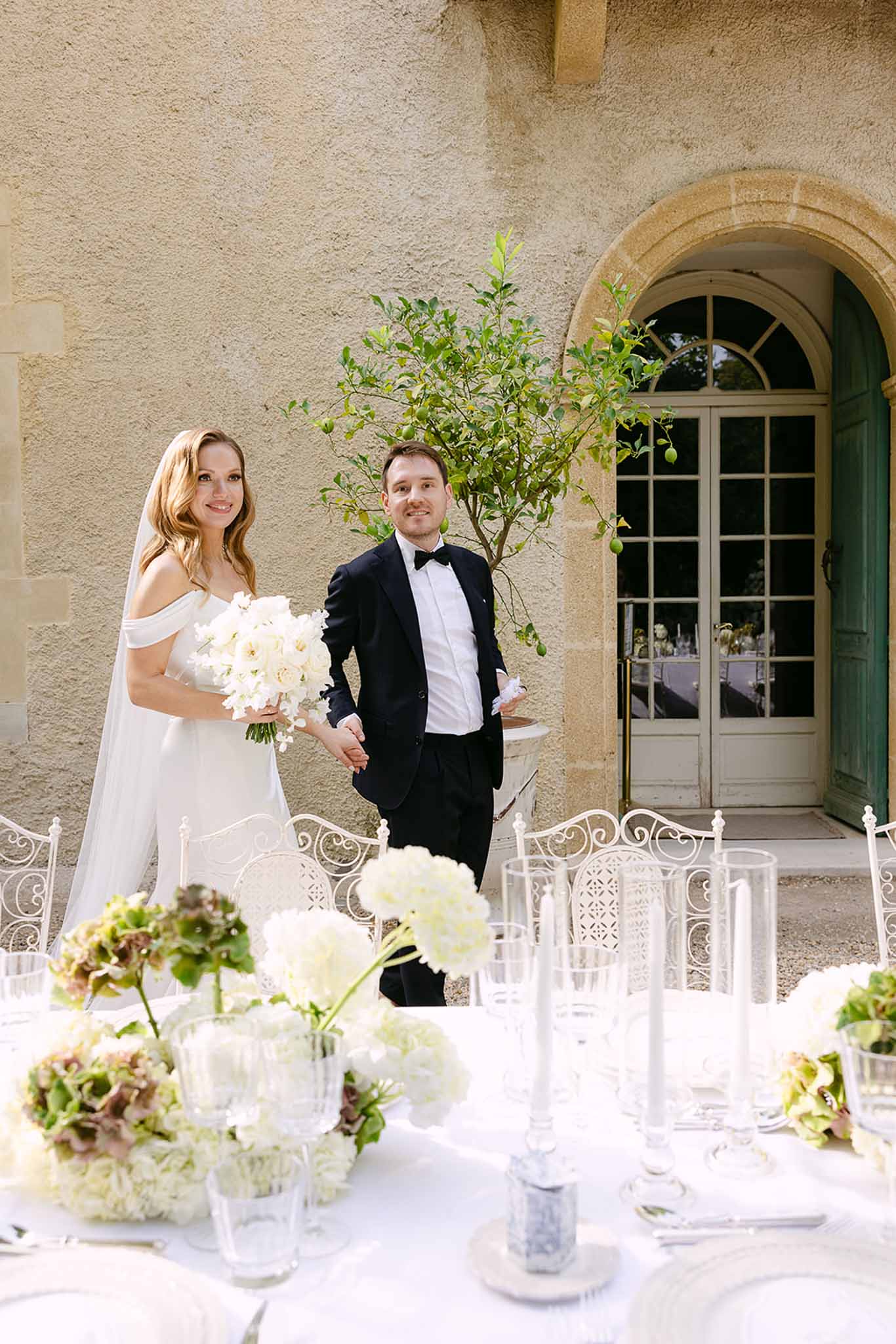Bride and groom smiling before chateau with white hydrangea reception table and taper candles in foreground
