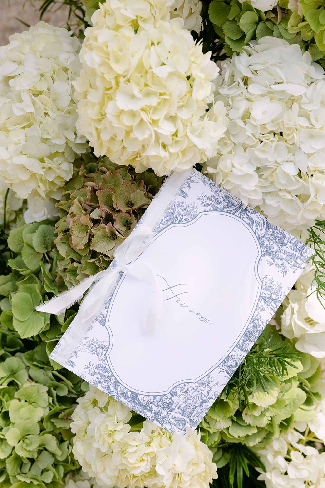 Toile de Jouy ceremony booklet with silk ribbon on white and sage hydrangea bed with rosemary sprigs