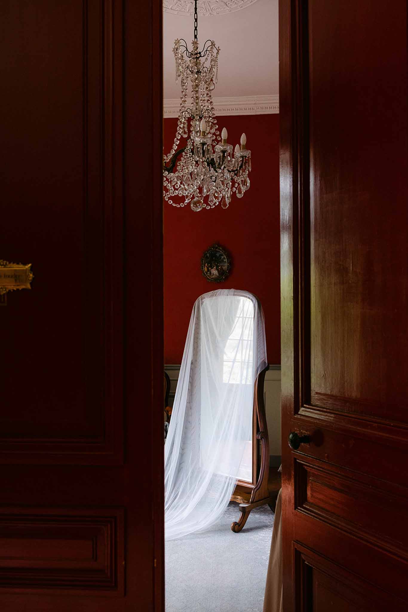 White bridal veil draped on antique cheval mirror in red-walled chateau room framed through mahogany doors