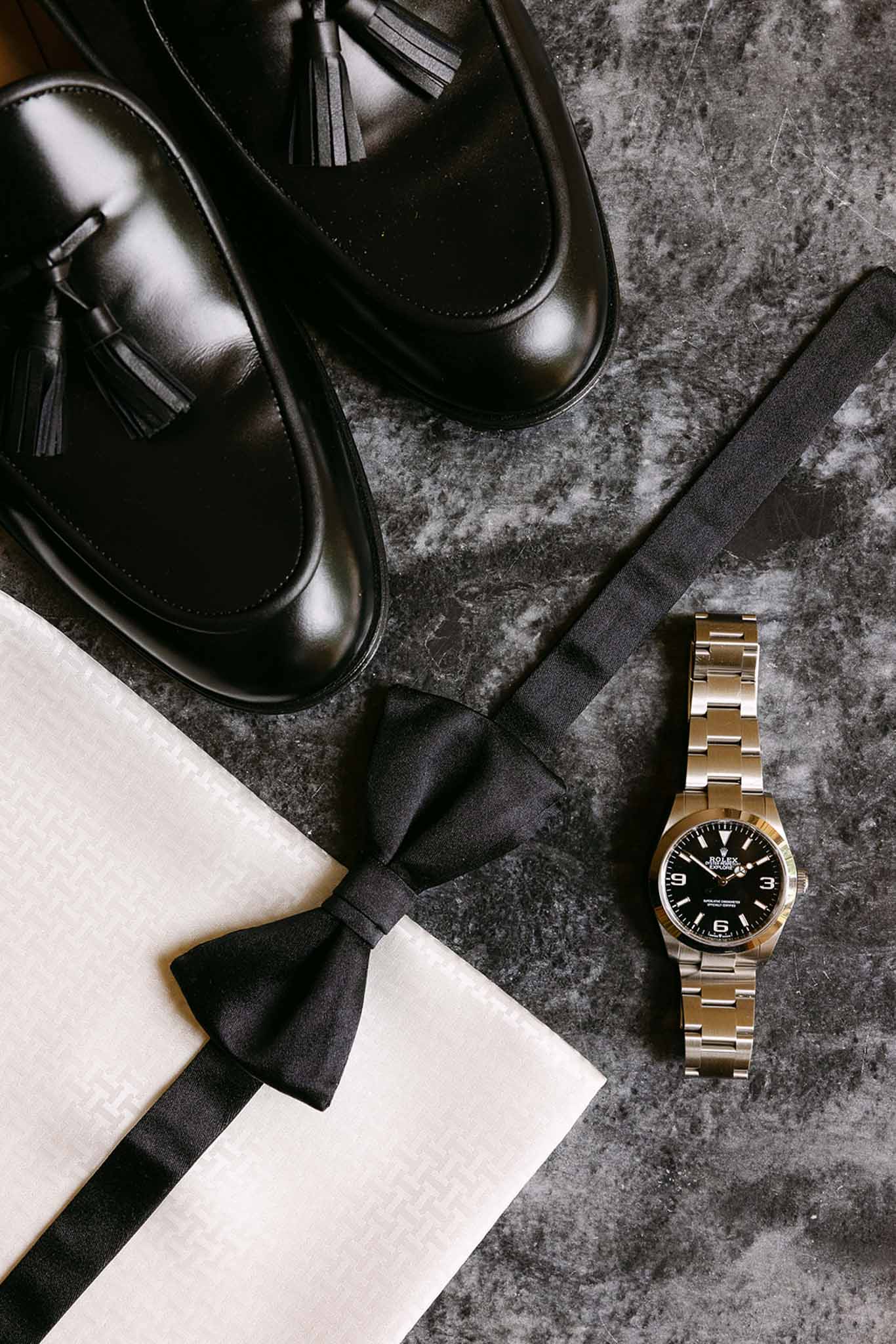 Groom flat lay on dark marble with black tassel loafers, satin bow tie, ivory pocket square, and Rolex watch