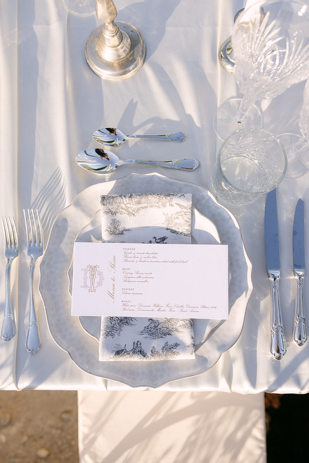 Place setting with toile de Jouy napkin, monogram menu card, crystal tumbler, and silver candelabra