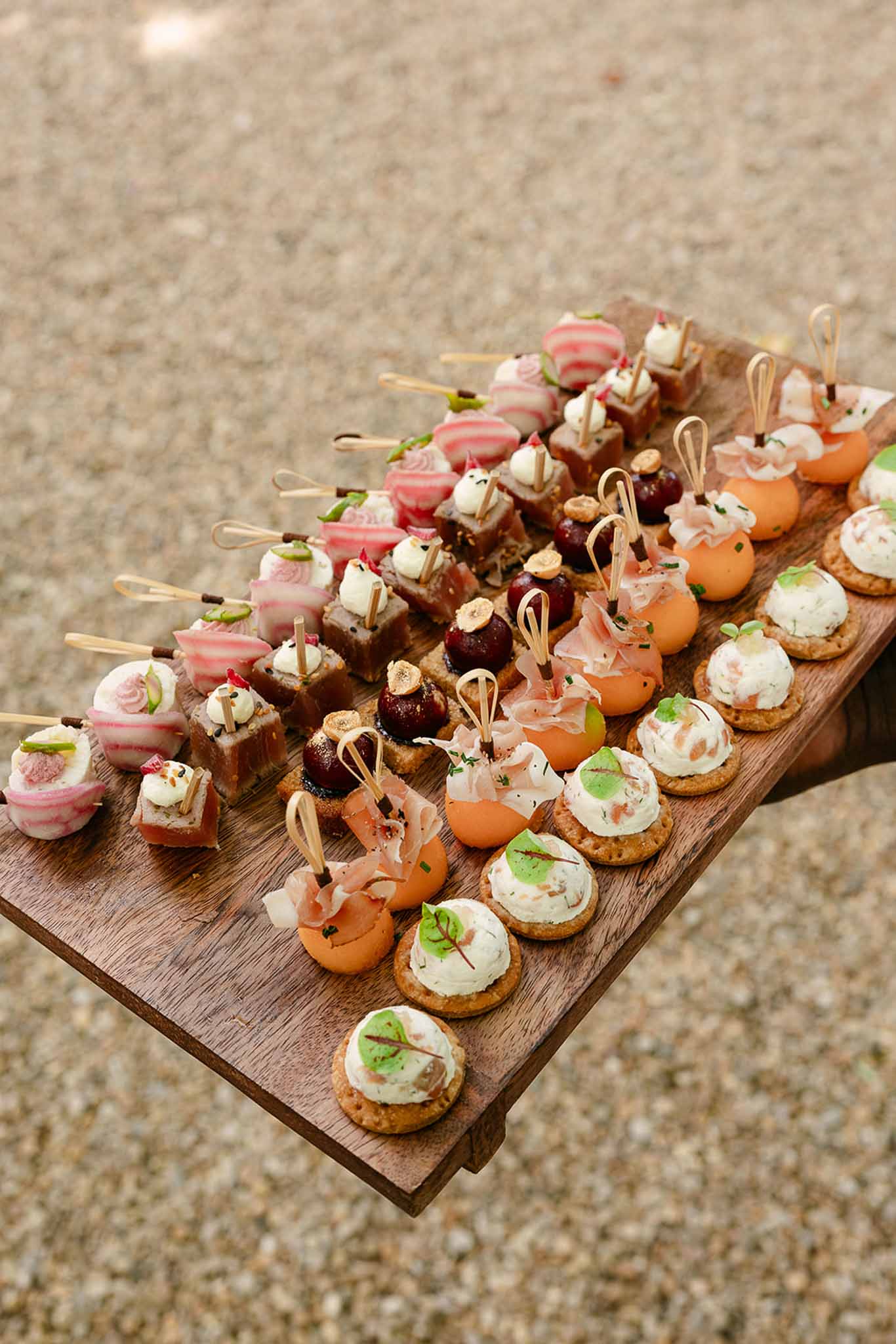 Wooden board with four rows of bite-sized canapes including melon prosciutto and herb cream cheese crackers