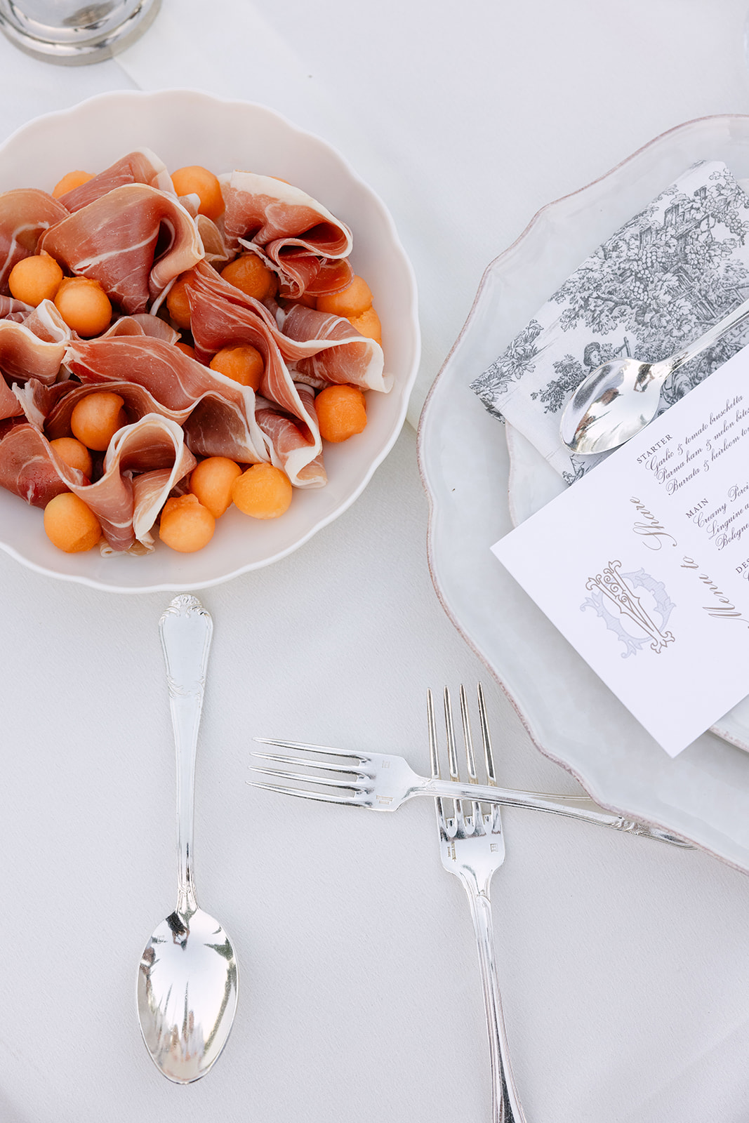 Place setting with melon and prosciutto starter, toile-illustrated menu card, and ornate silver cutlery