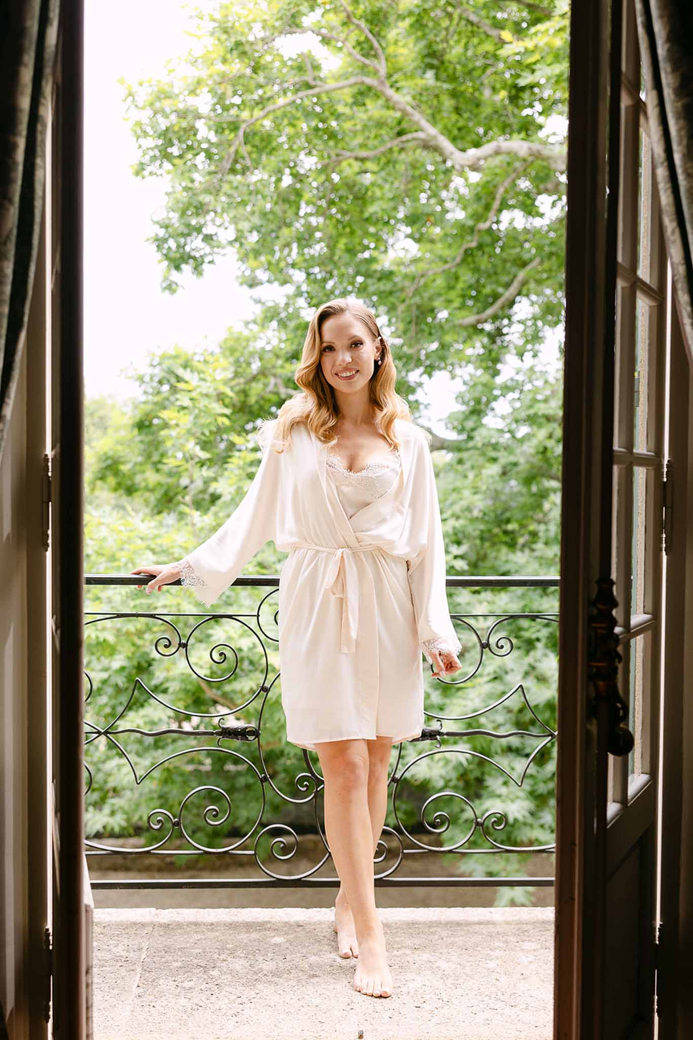 Bride in blush silk robe standing barefoot on a balcony framed by tall French doors with wrought-iron railings