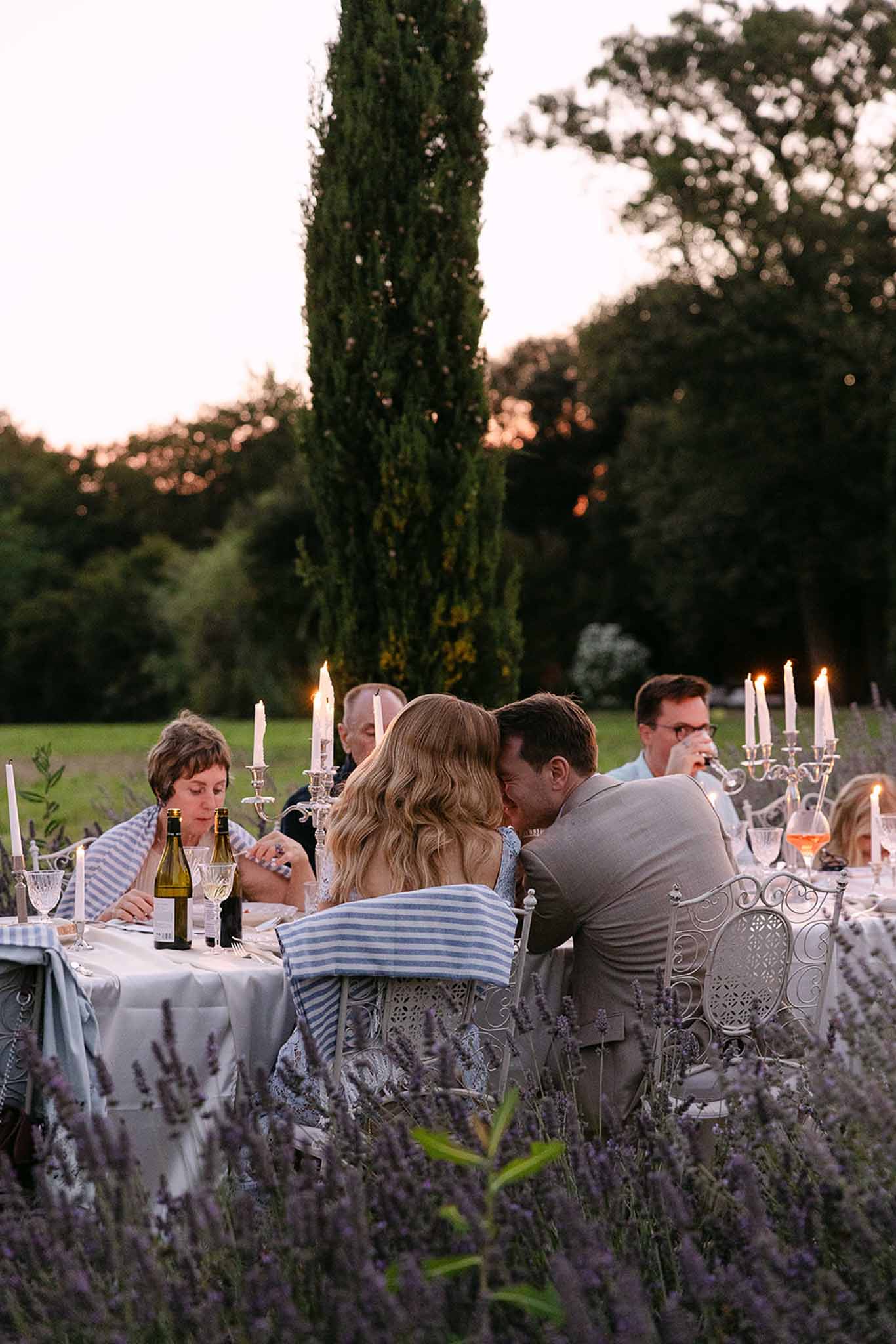 Dusk garden dinner with lavender border white candelabras and wrought-iron chairs beneath cypress tree