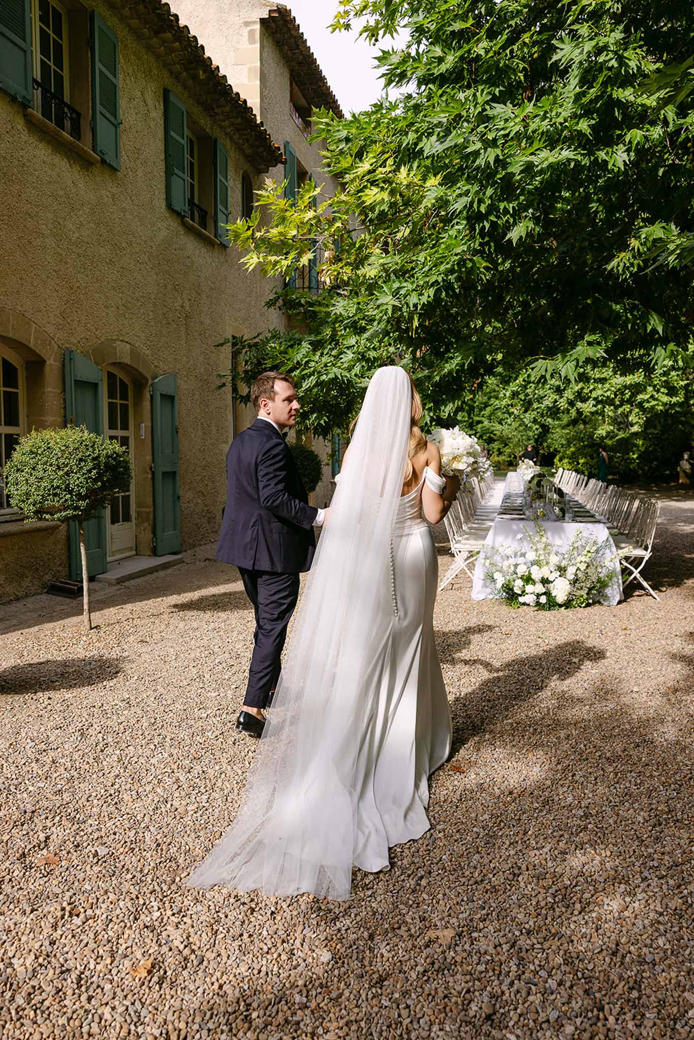 Bride and groom from behind walking toward white-draped reception table at teal-shuttered Provencal mas