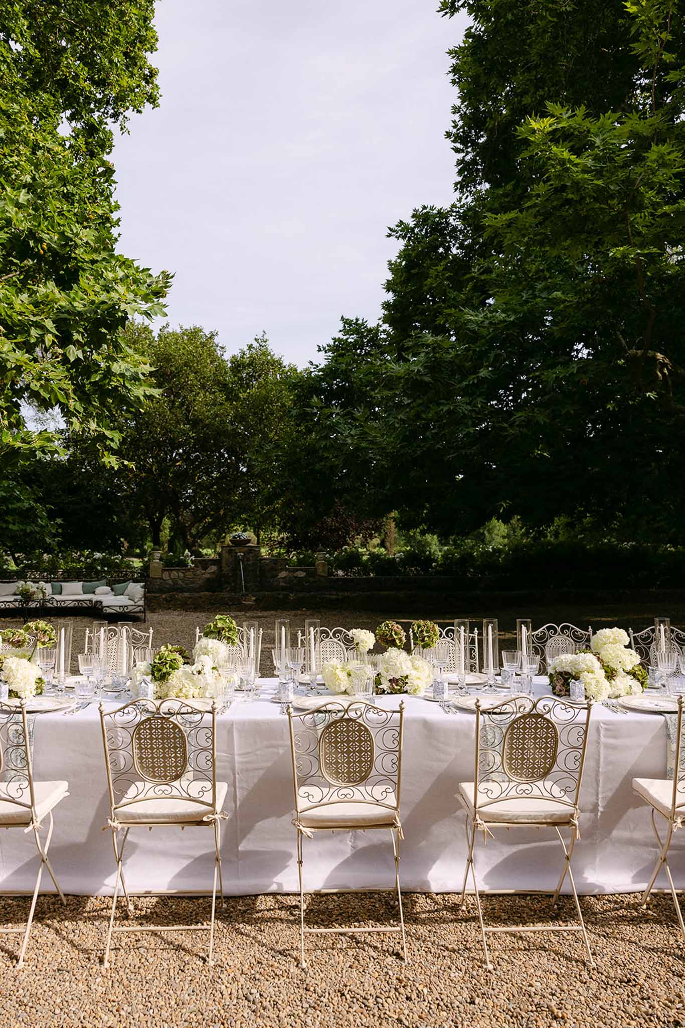 Long table with white and green hydrangea clusters and wrought-iron lattice chairs on gravel under trees