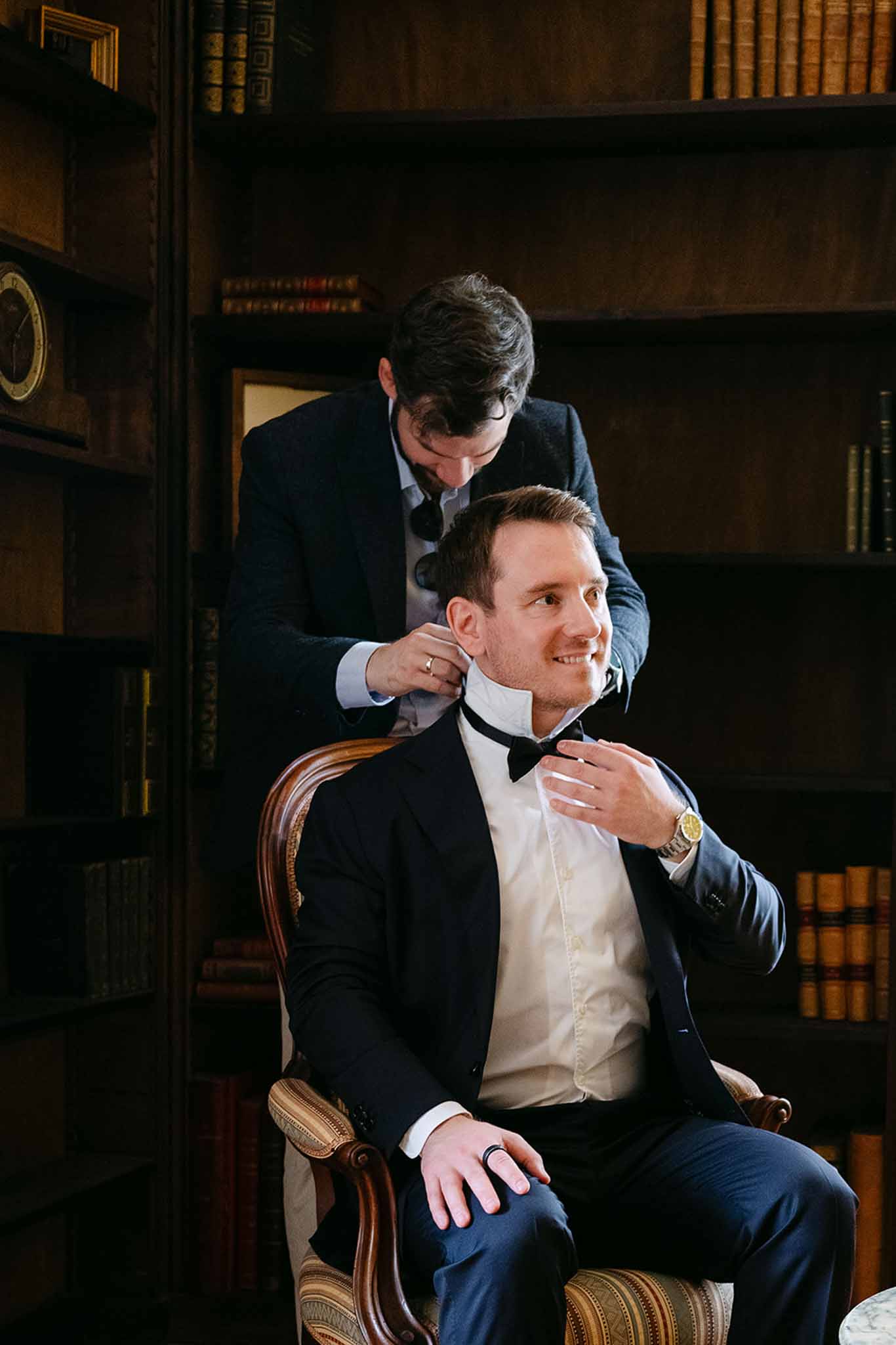 Groomsman adjusting groom bow tie in library with leather-bound bookshelves
