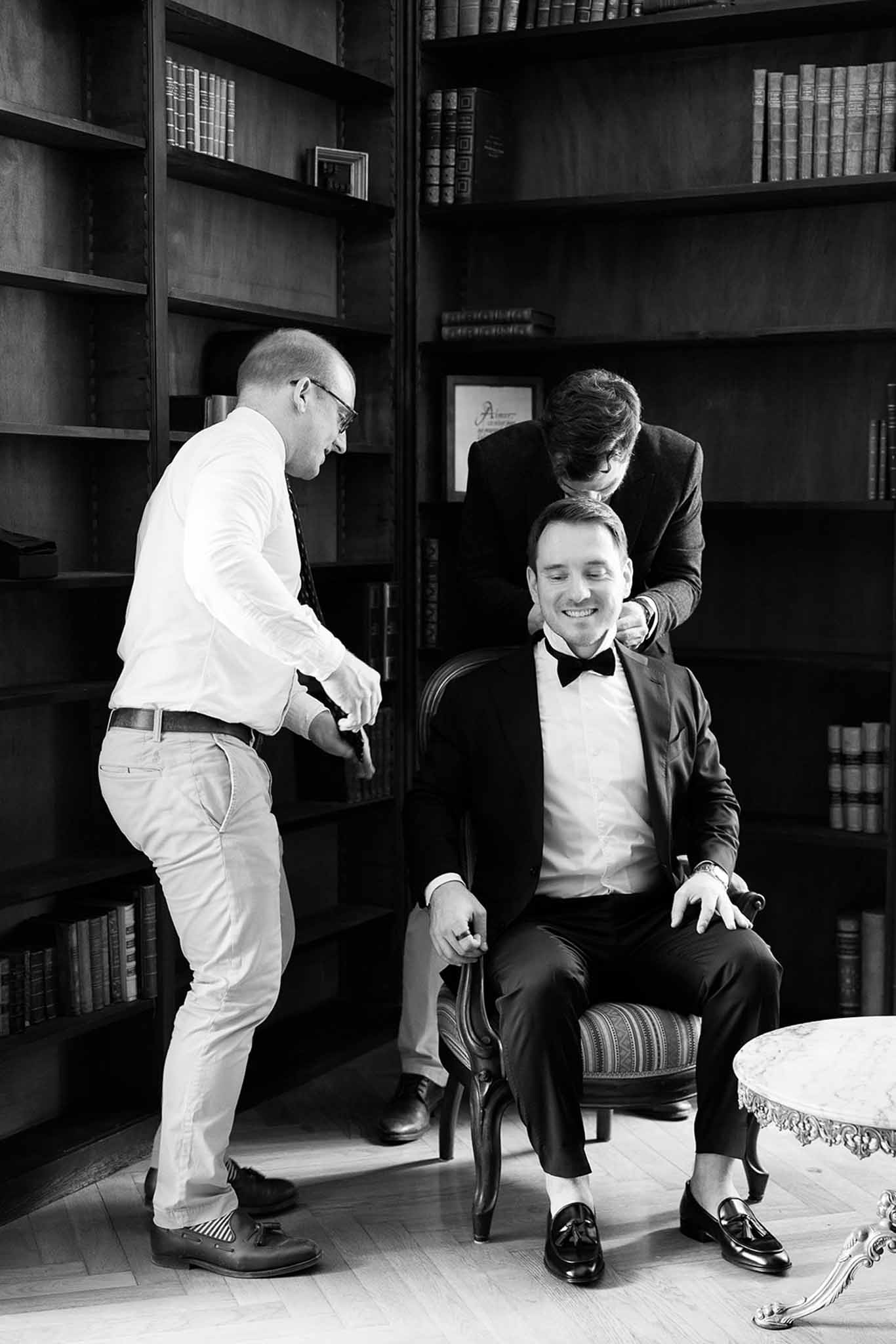 Black and white groom in tuxedo being helped by two groomsmen in wood-panelled library