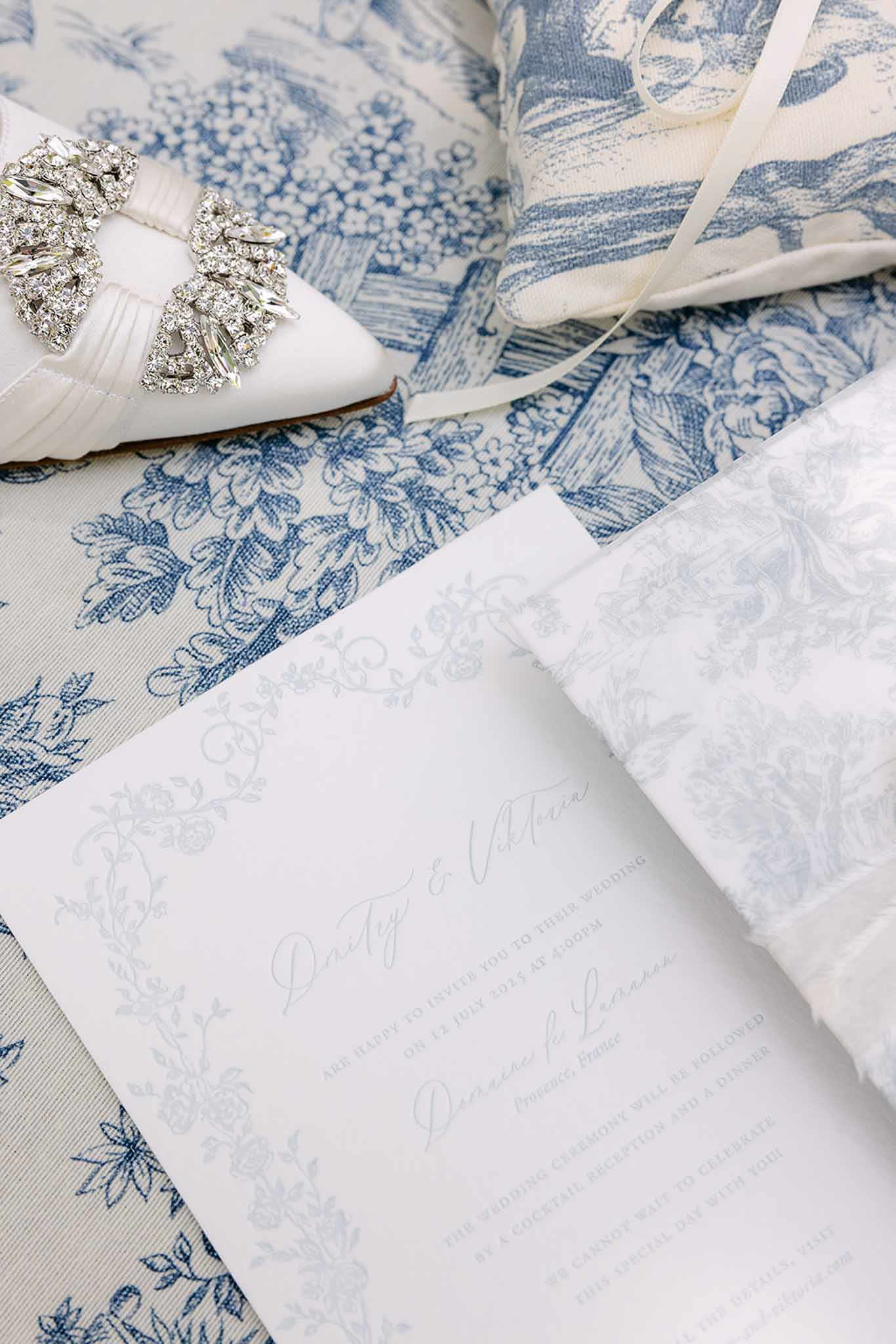 Wedding invitation with blue botanical border on toile fabric beside crystal-embellished white satin bridal shoe