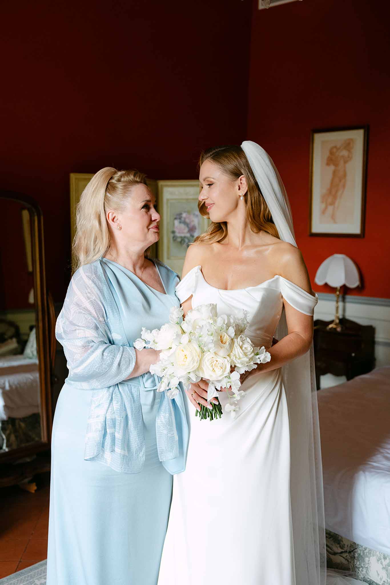 Bride in ivory off-shoulder satin gown with mother in powder blue dress both touching ivory rose bouquet in red-walled room