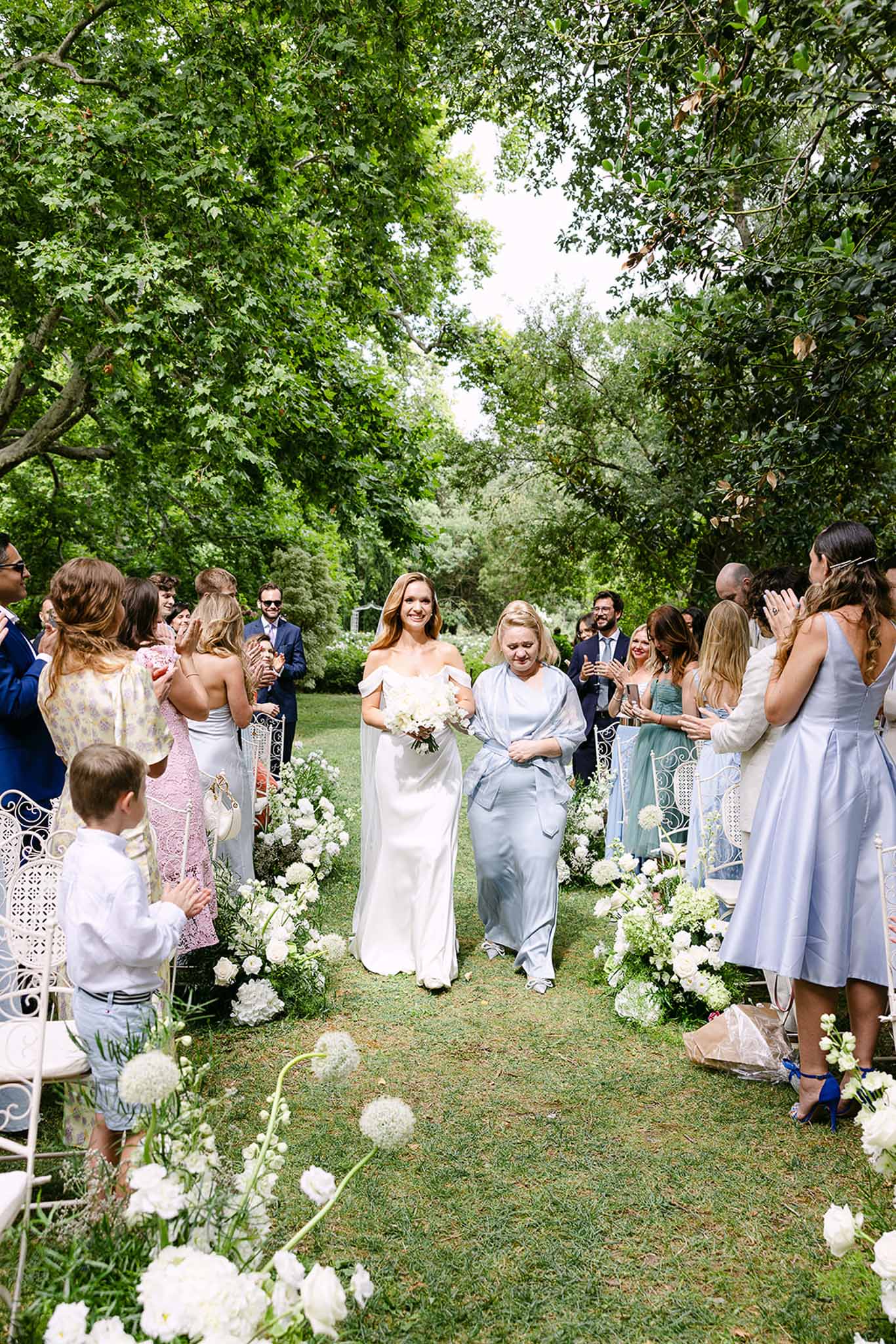 Bride in white satin gown with cape walks aisle with mother past all-white peony and allium arrangements