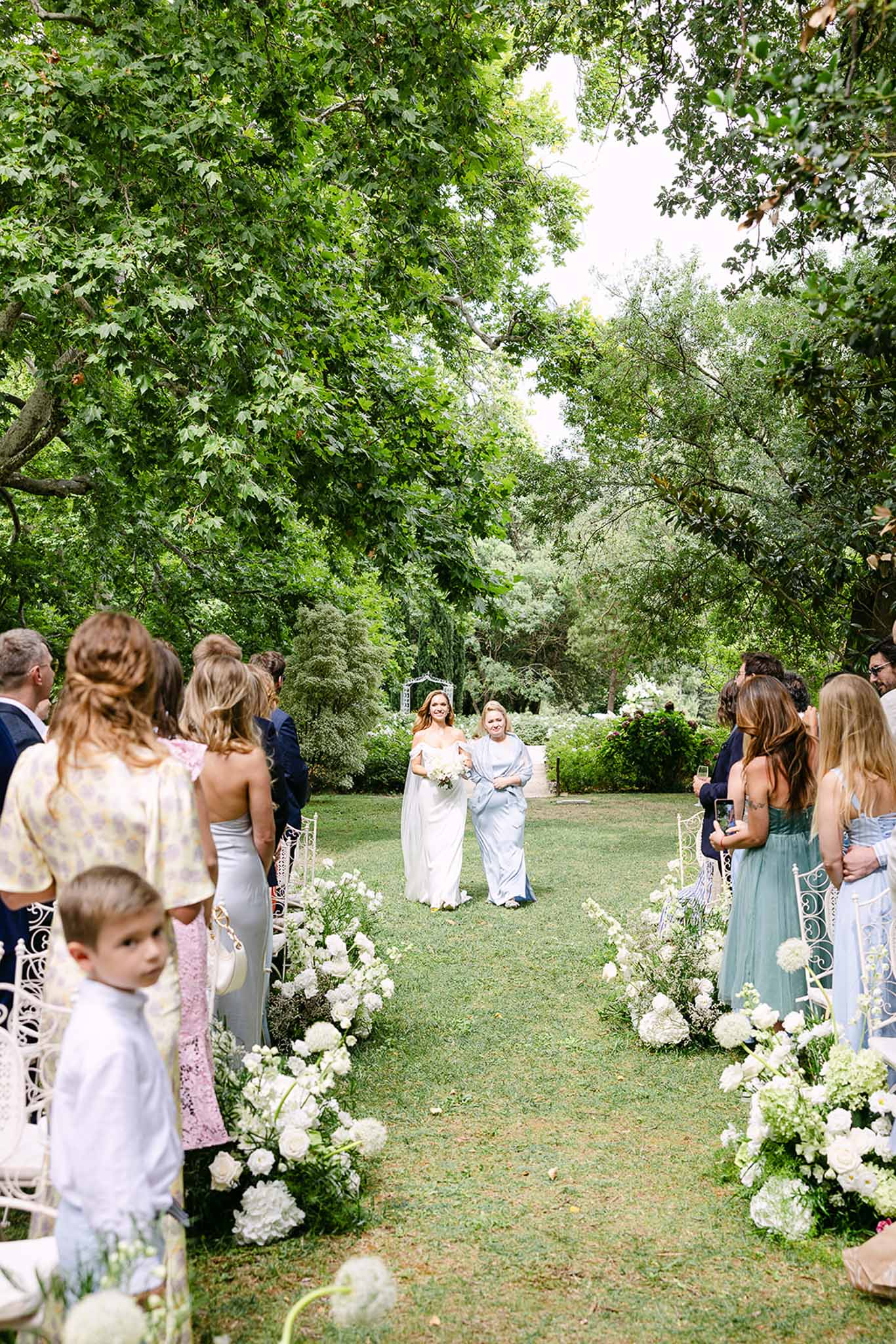 Bride walking down aisle with companion past white peony and rose ground arrangements and wrought-iron chairs on lawn