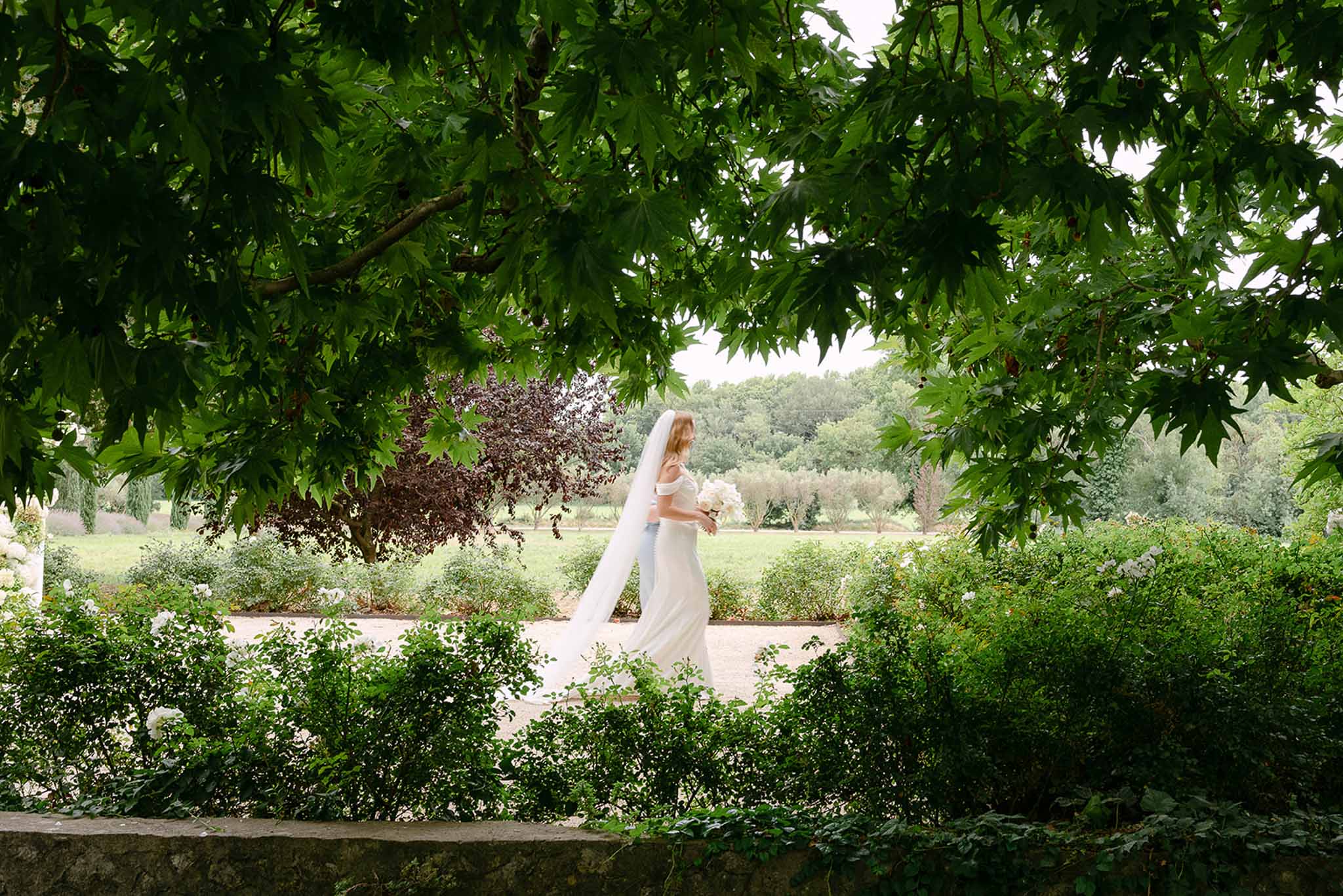 Bride walking along gravel path in ivory off-shoulder gown with cathedral veil framed by garden hedgerows and trees