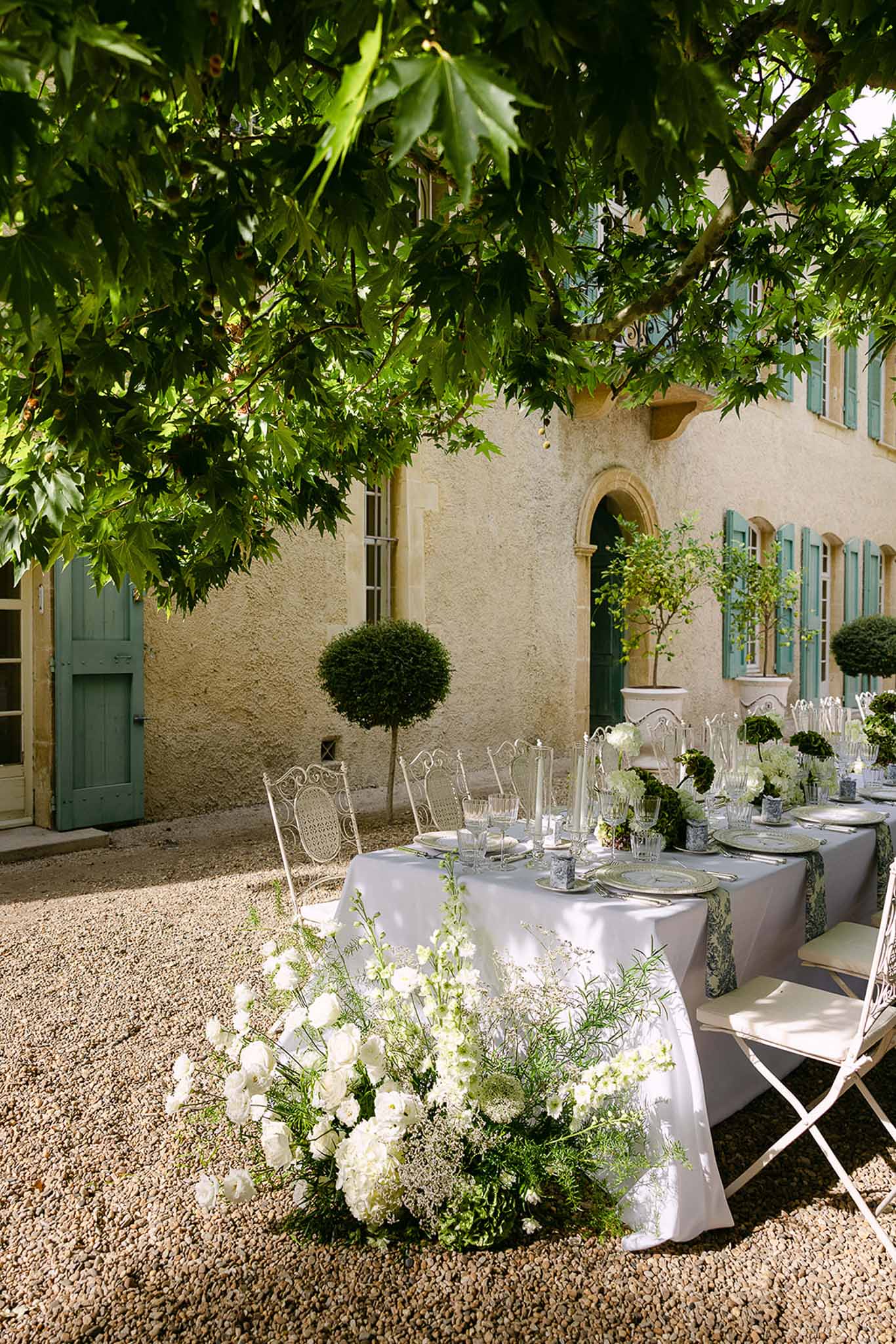 Reception table in Provencal gravel courtyard with botanical runner, white ranunculus, and wrought-iron chairs