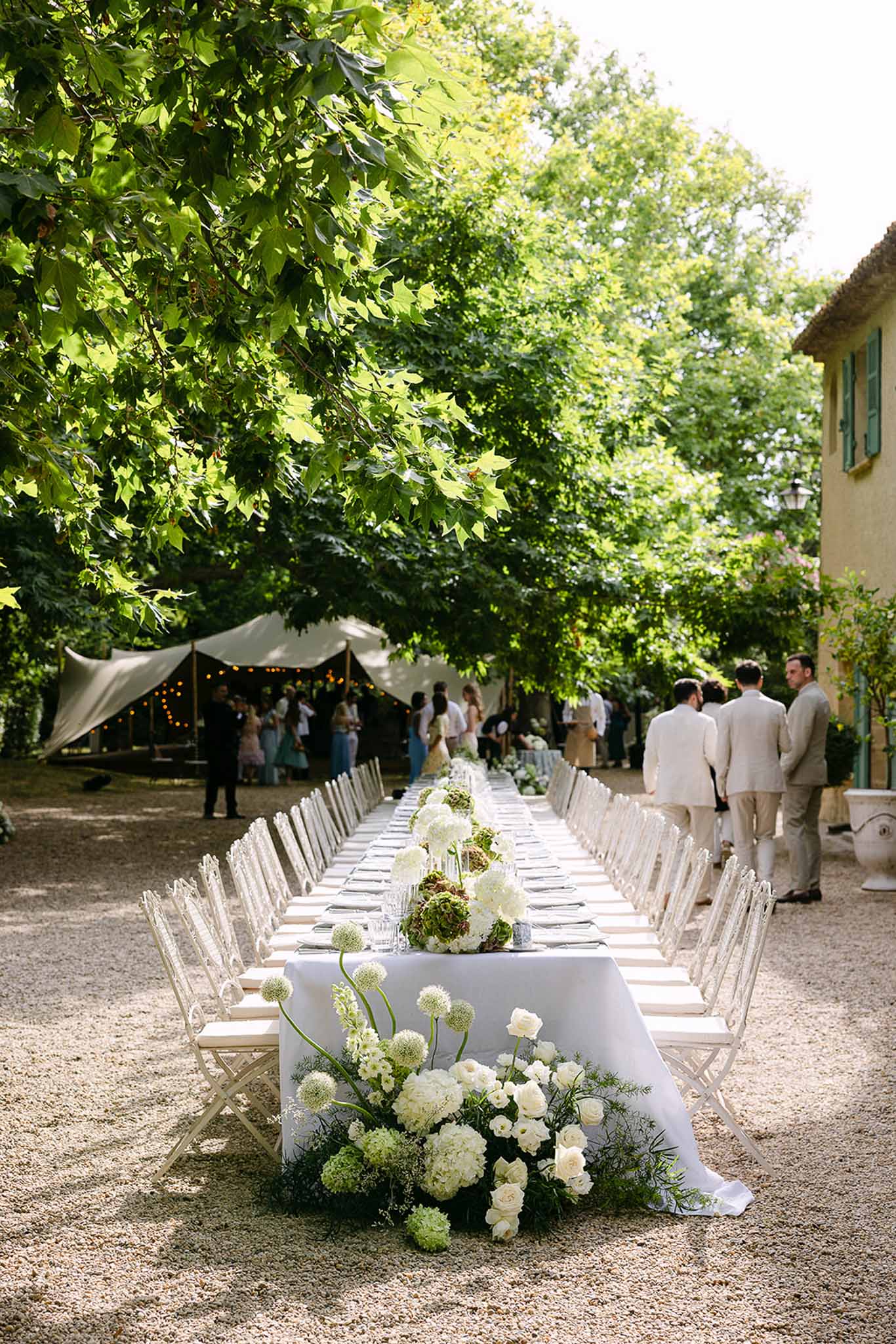 Long feasting table with white hydrangea and allium runner white chairs tent with fairy lights and Provencal stone building