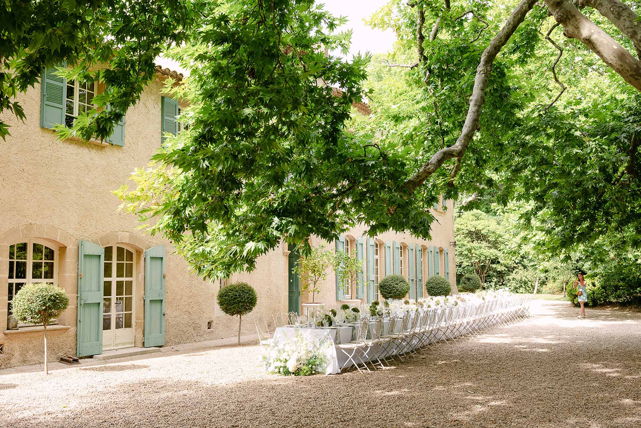 Long banquet table with white linens and floral centerpieces along honey-toned Provencal mas courtyard