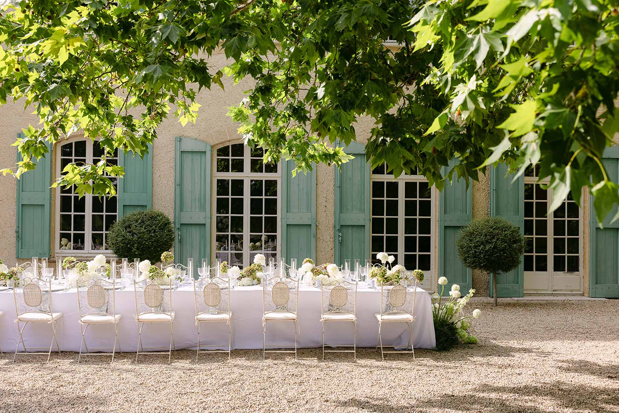 Long reception table with white hydrangea centerpieces and bistro chairs on gravel courtyard of Provencal manor