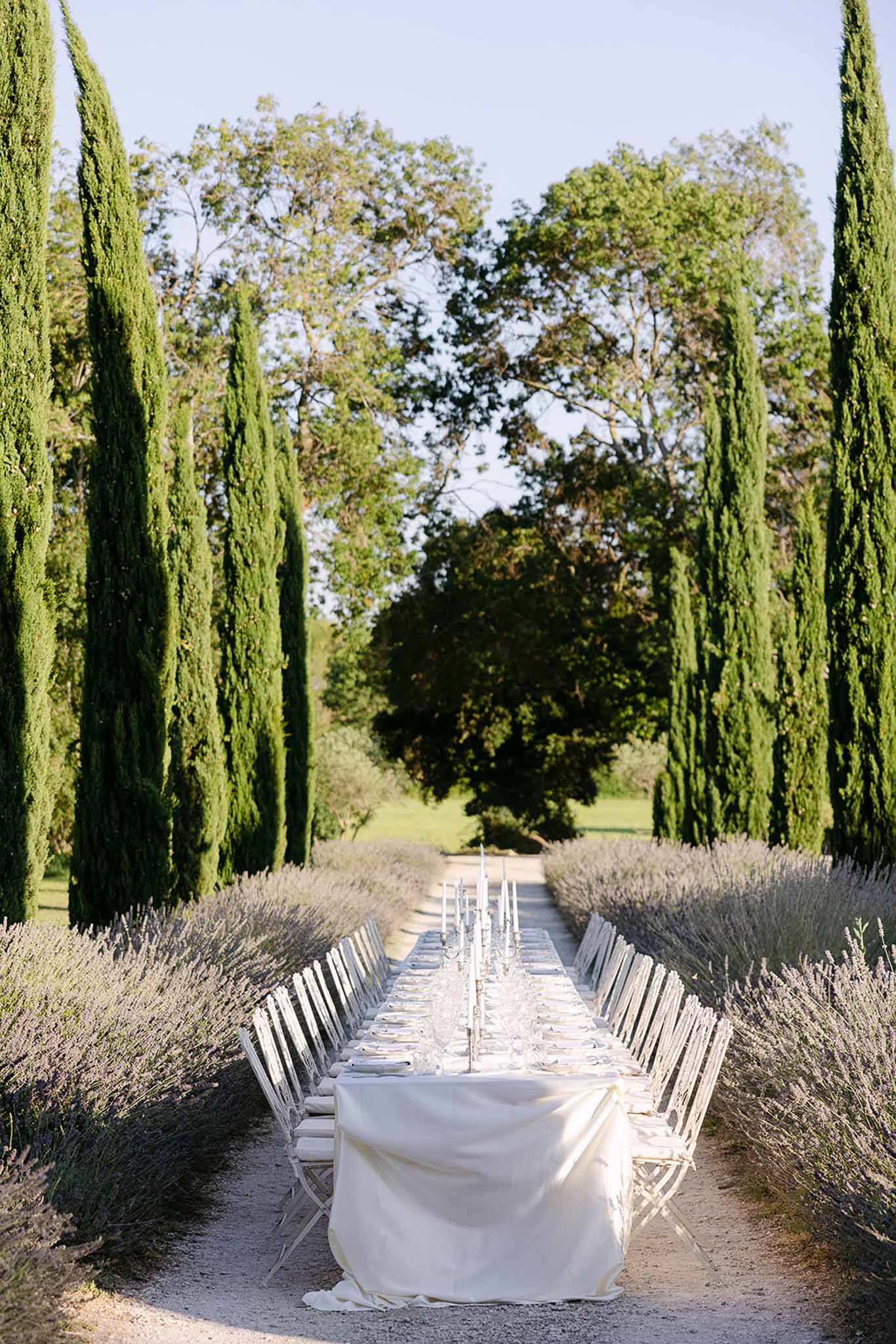 All-white feasting table with silver candelabras between lavender hedges and cypress trees on gravel allee