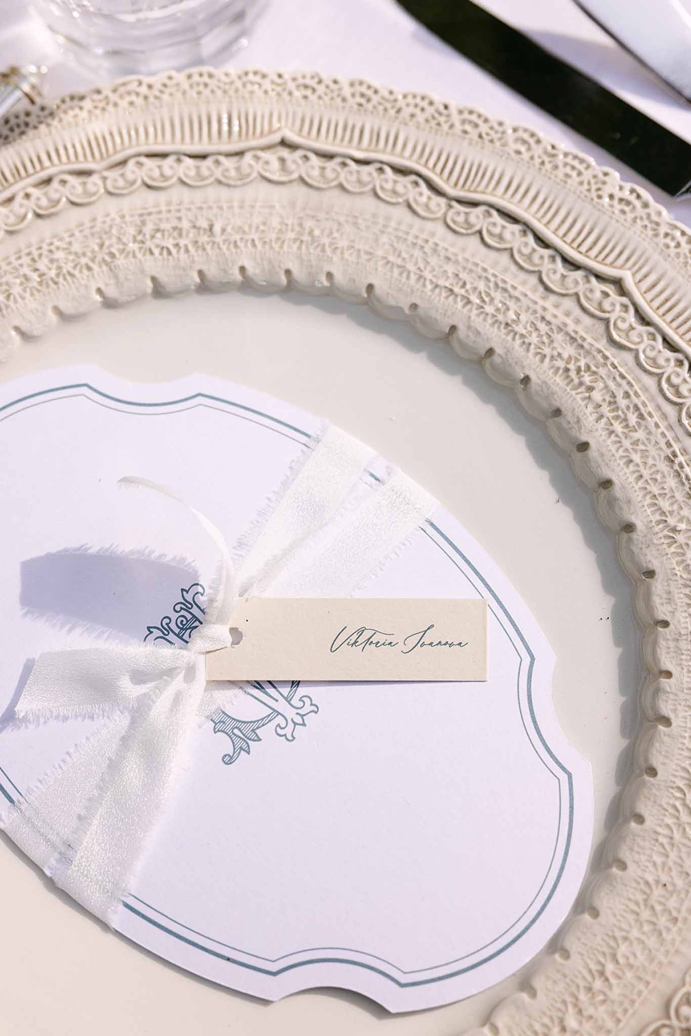 Wedding place setting with ivory embossed charger plate, shield-shaped menu card with dusty blue monogram, and satin name tag