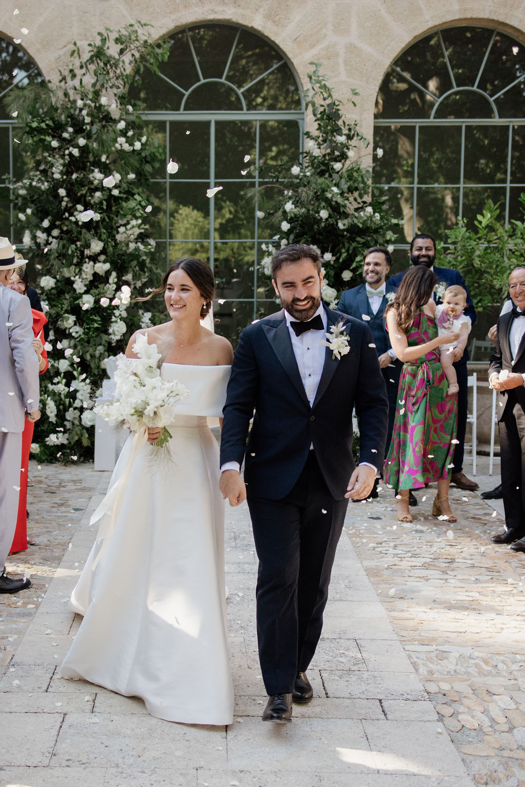 Couple walking up aisle as guests throw white petals, with floral columns framing stone courtyard ceremony backdrop