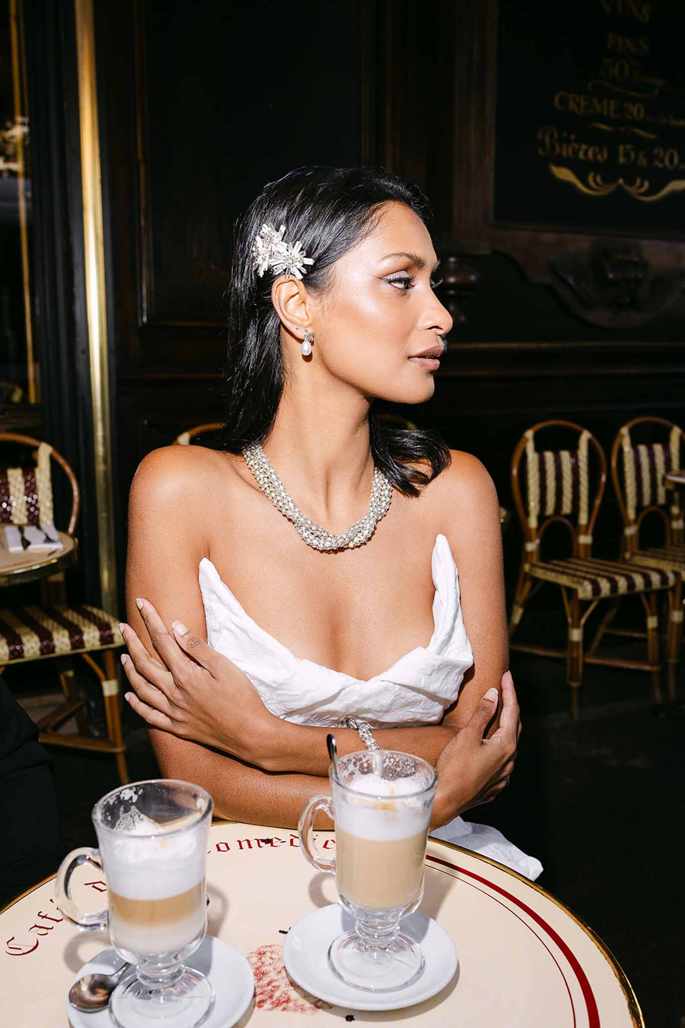 Bride in white strapless bodice and crystal necklace seated at a Parisian cafe with latte drinks