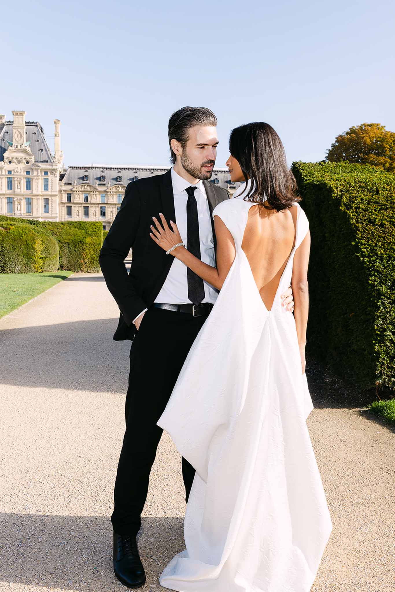 Bride and groom face each other in Tuileries Garden with Louvre in background, bride in open-back white gown