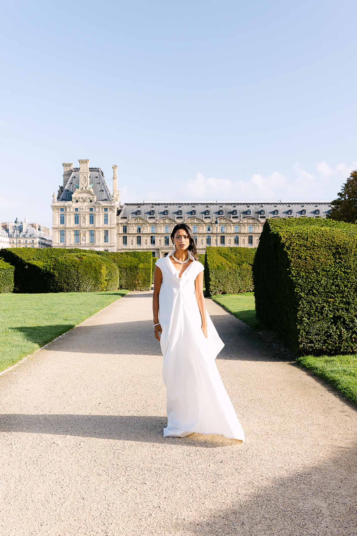 Bride in minimalist white V-neck gown with pearl choker standing in Jardin des Tuileries with Louvre behind