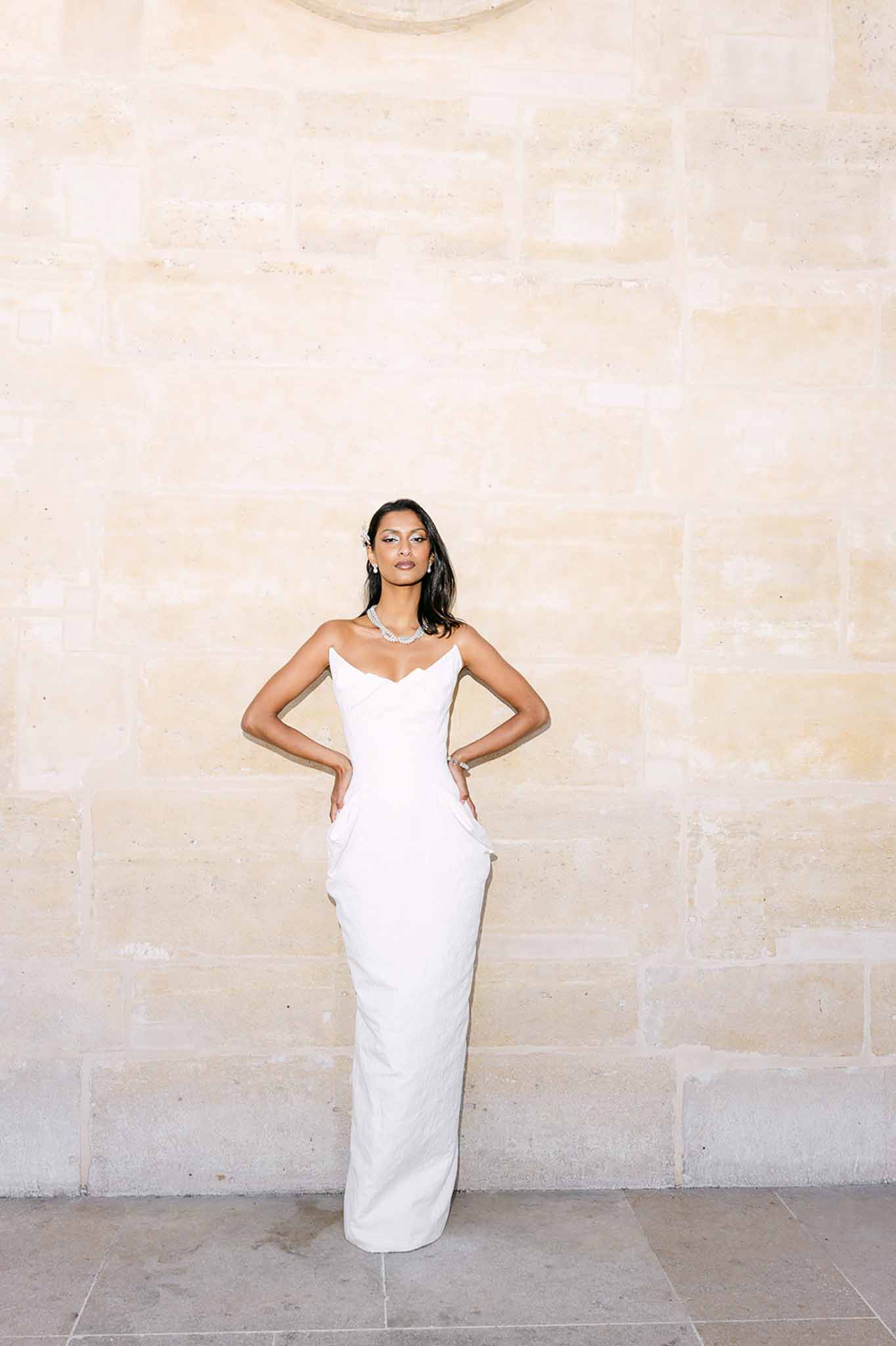 Bride in strapless white column gown with crystal necklace standing against limestone wall