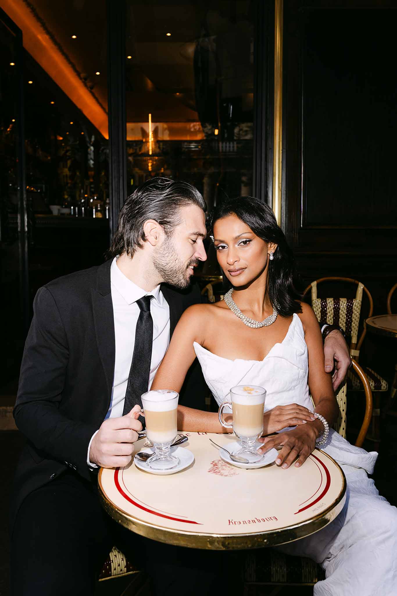 Couple at Parisian brasserie table with lattes bride in strapless gown with crystal necklace groom in dark suit