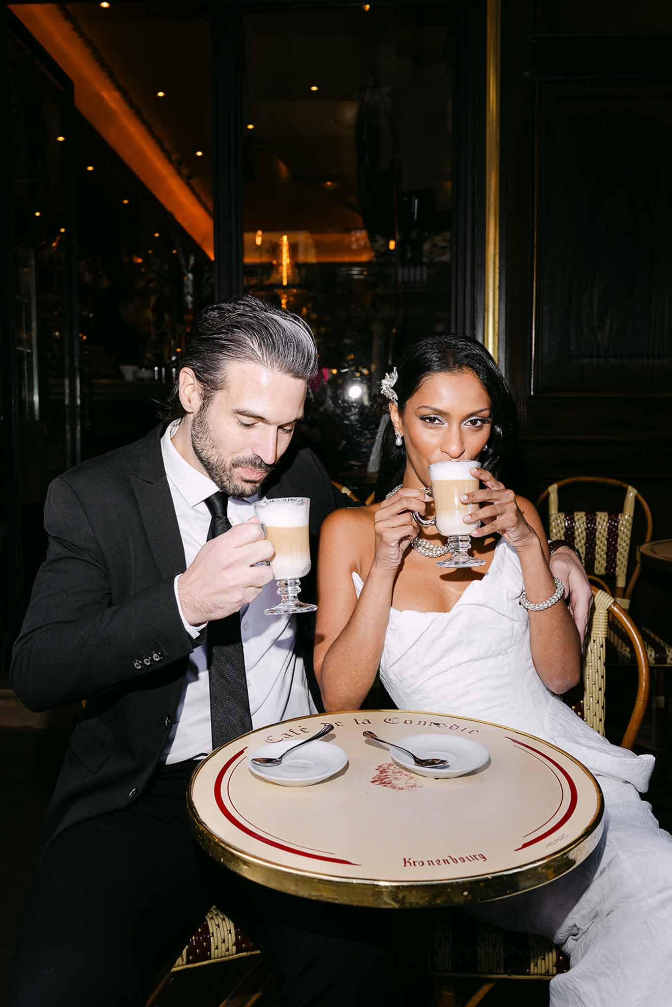 Bride and groom sitting at a Parisian cafe table drinking latte macchiatos at night
