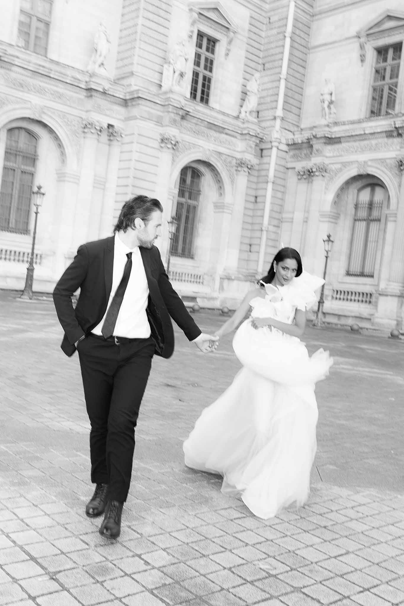 Black and white motion-blurred couple running on Paris cobblestone courtyard in ruffled tulle gown