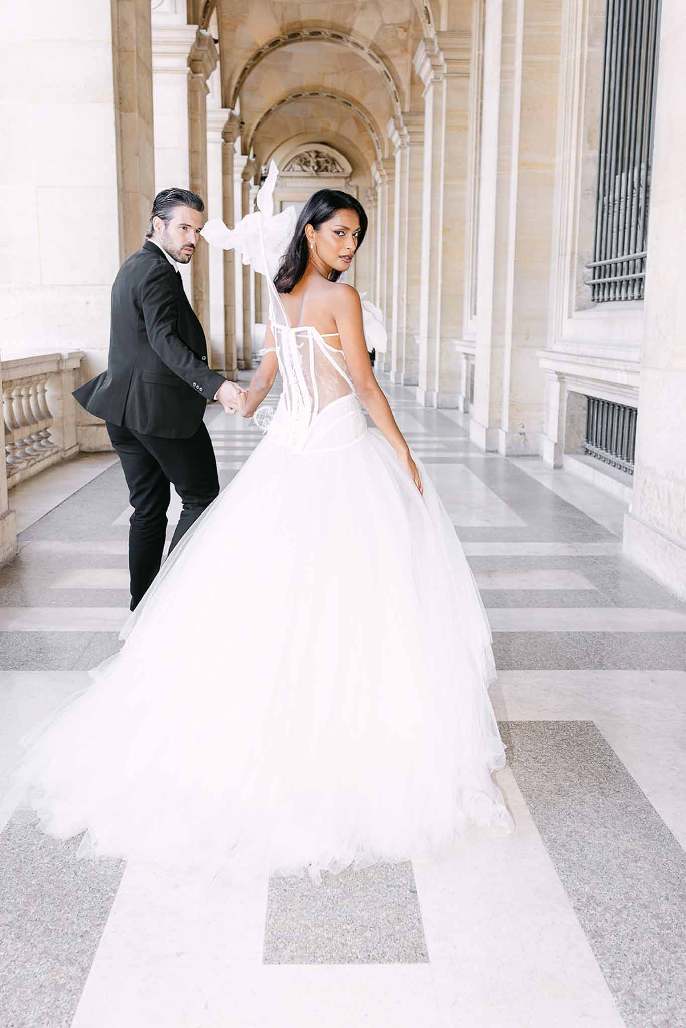 Bride in strapless ball gown with sculptural headpiece and groom in black suit walking through Parisian colonnade