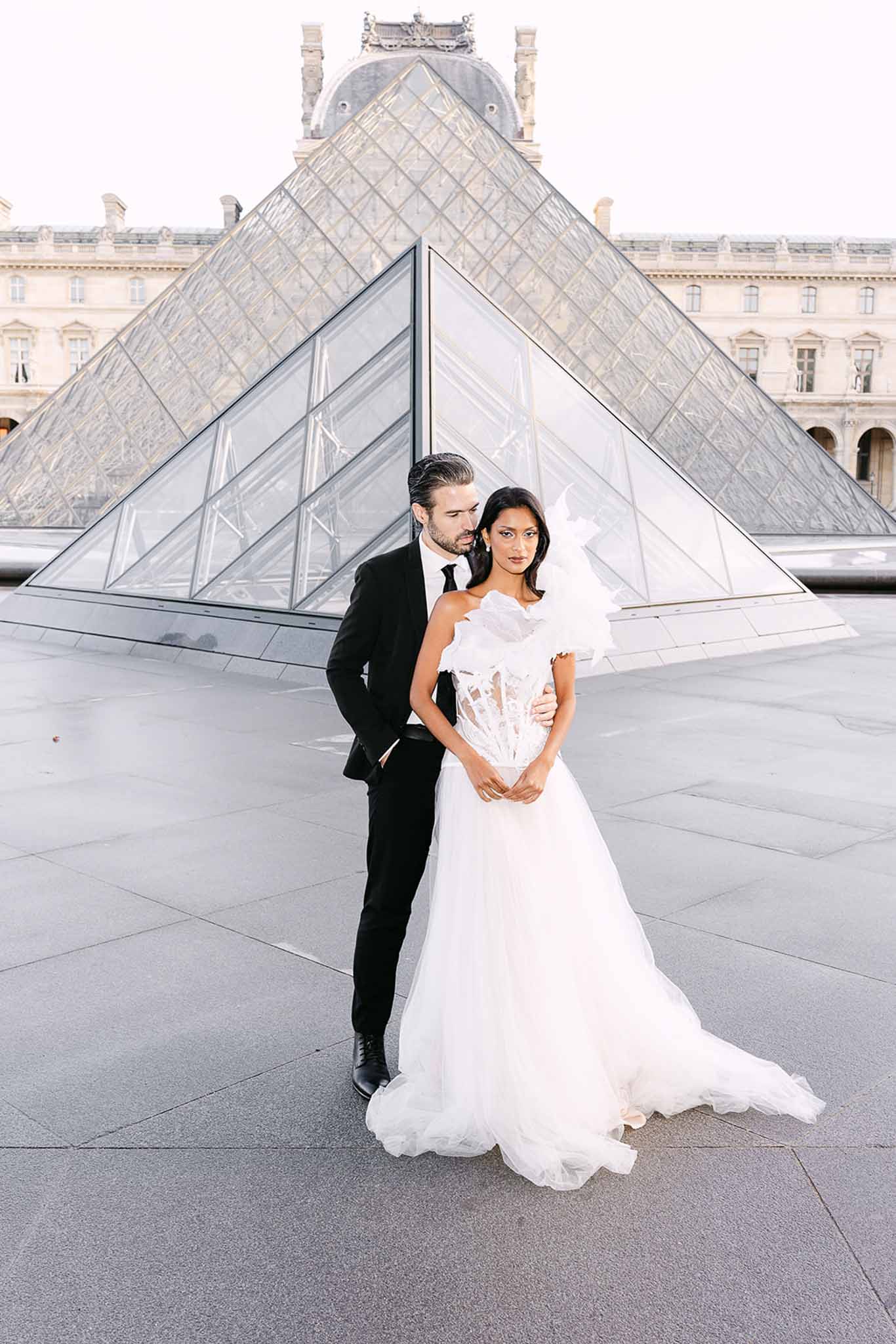 Bride in white ballgown and groom in black suit posing at the Louvre pyramid in Paris