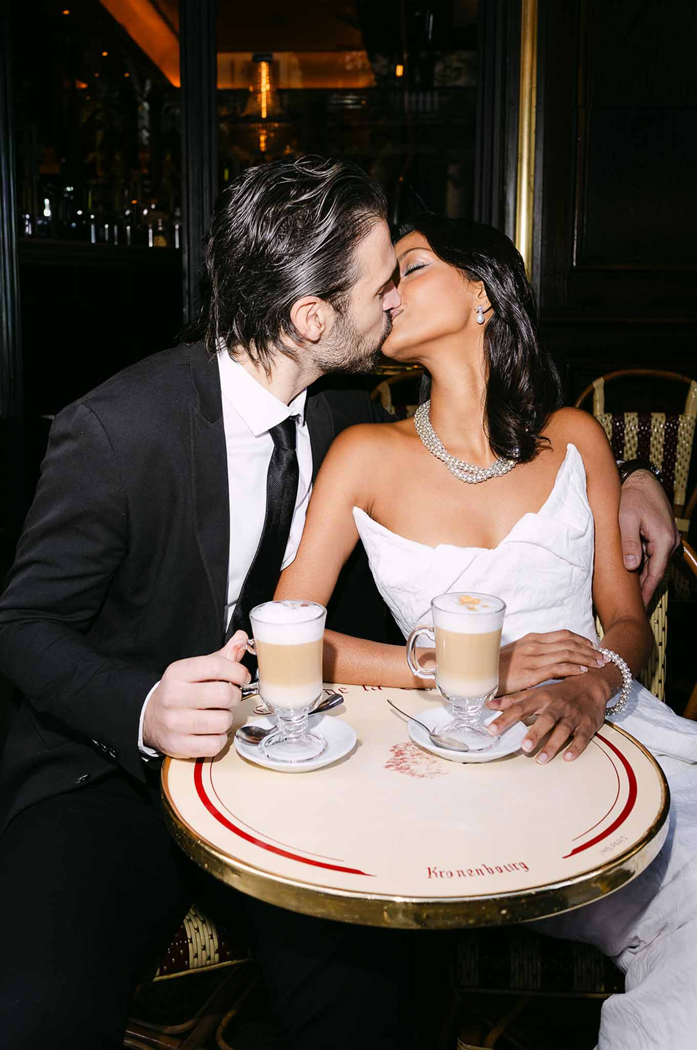 Bride and groom kissing at a Parisian cafe table with latte drinks in warm ambient lighting