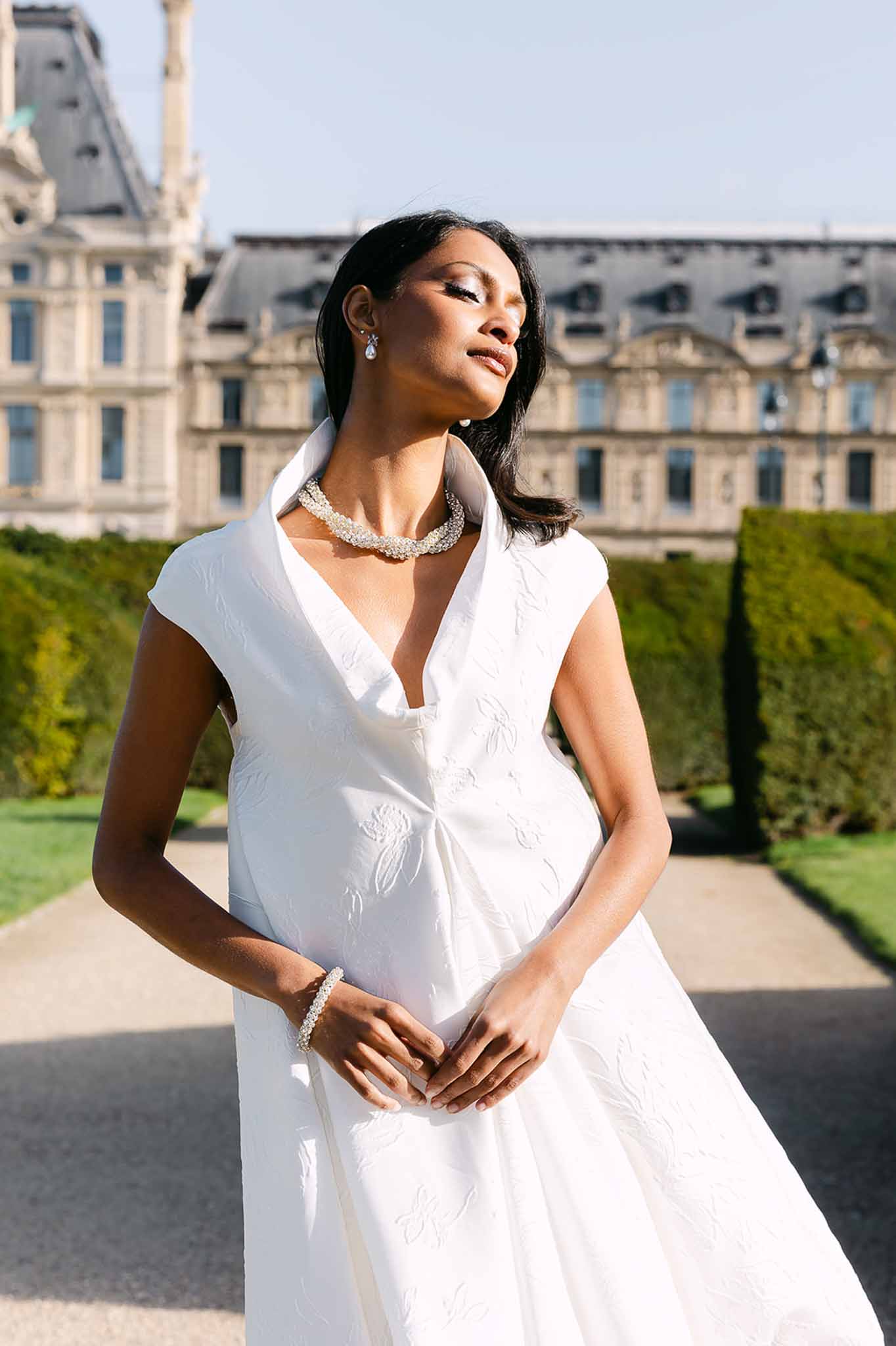Bride in white jacquard cap-sleeve dress with pearl choker necklace in French garden with Parisian building backdrop