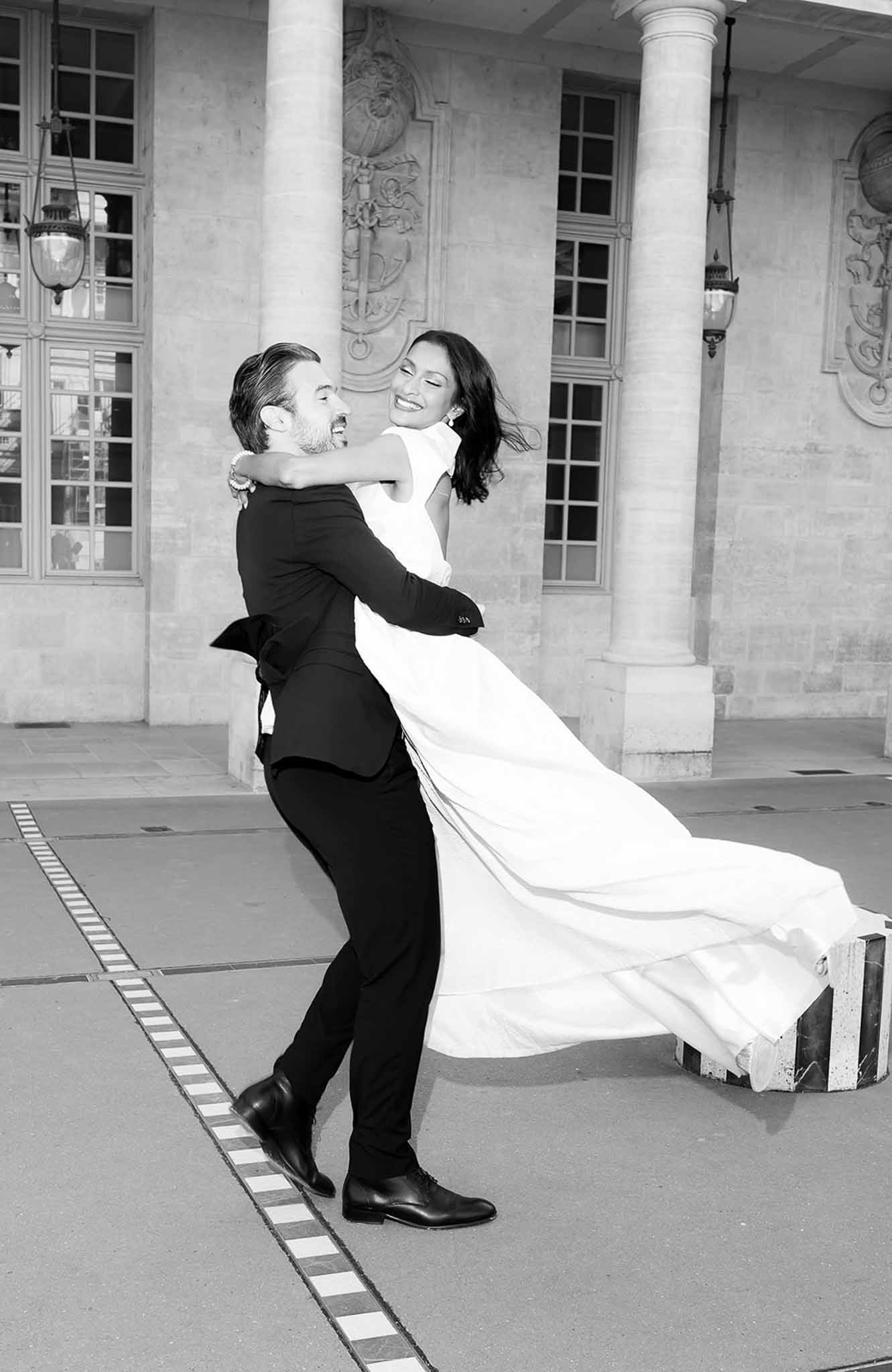 Black and white groom spinning bride in high-neck gown before Parisian stone columns and carved reliefs