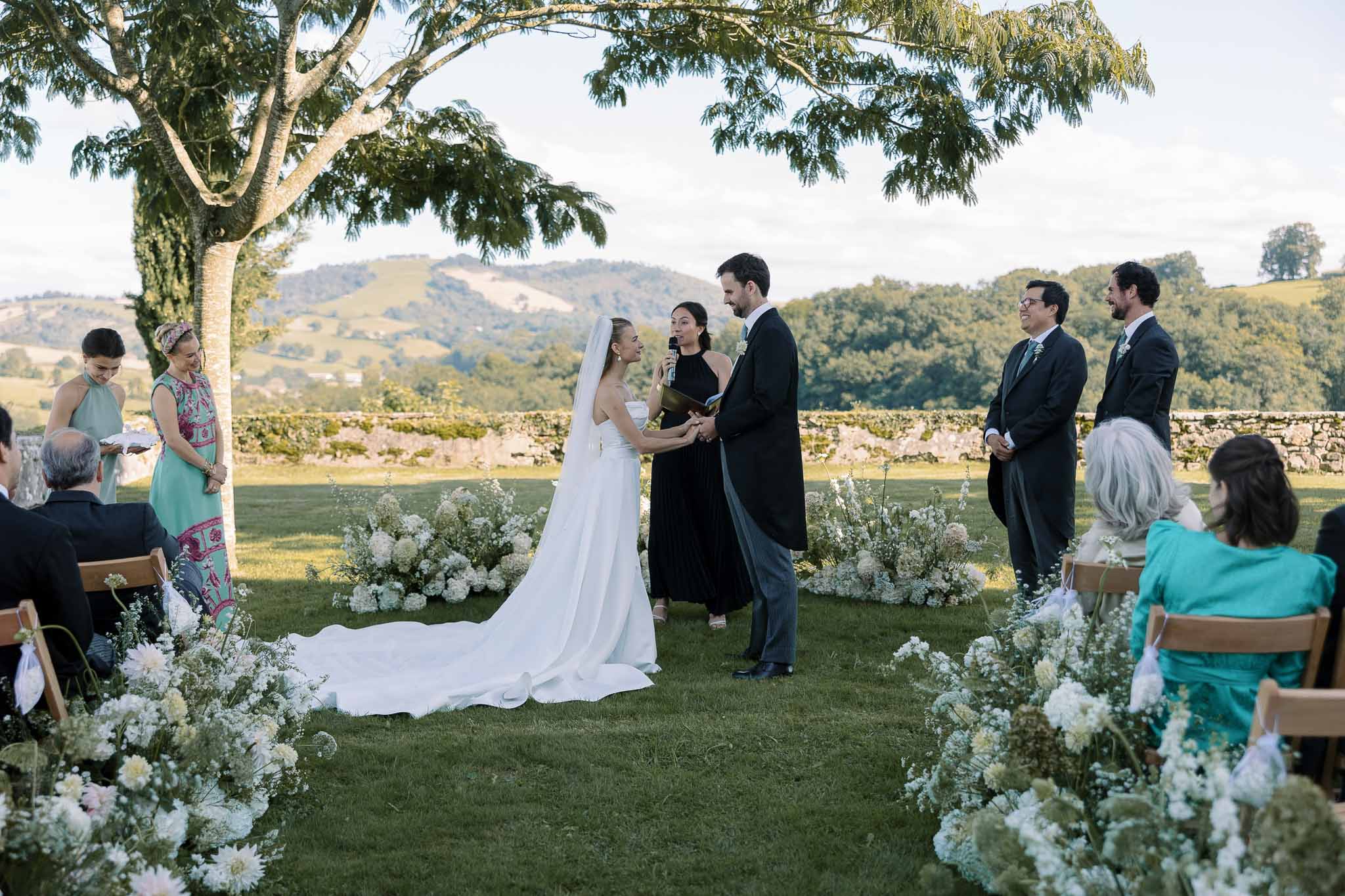 Outdoor wedding ceremony on a hilltop lawn with bride and groom exchanging vows among white floral arrangements