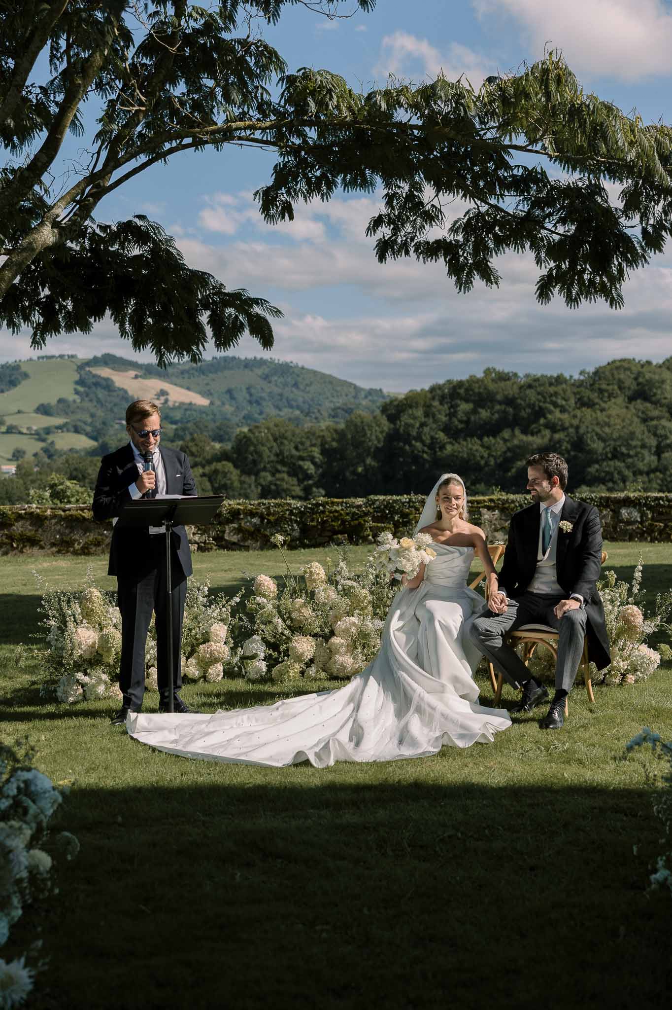 Seated couple at outdoor ceremony with white hydrangea ground installation and hedge backdrop