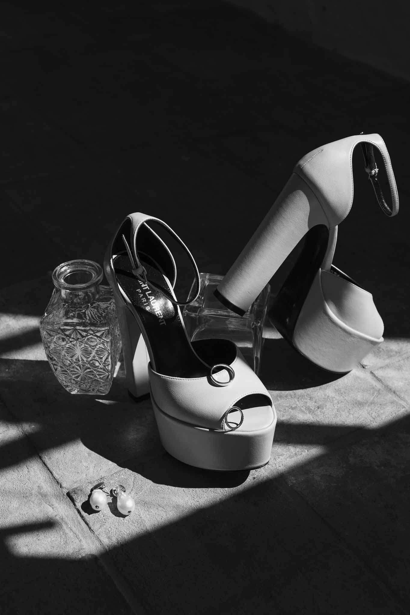Black and white flat lay of Saint Laurent white platform heels with wedding bands and pearl earrings