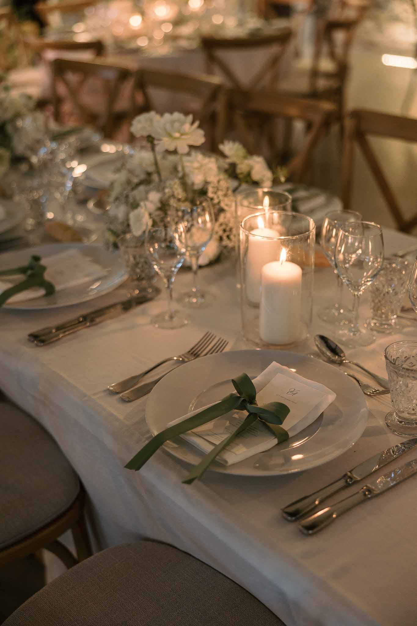 Candlelit reception table with white dahlia runner, sage green ribbon menus, and hurricane candle holders