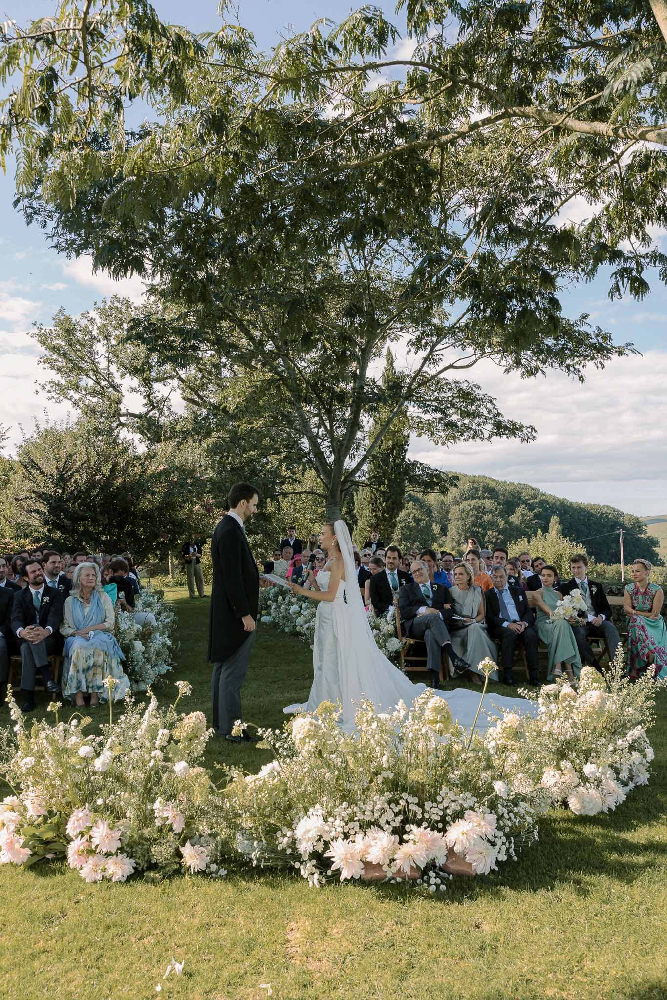 Outdoor ceremony with couple exchanging vows behind semicircular pink dahlia and white delphinium ground arrangement