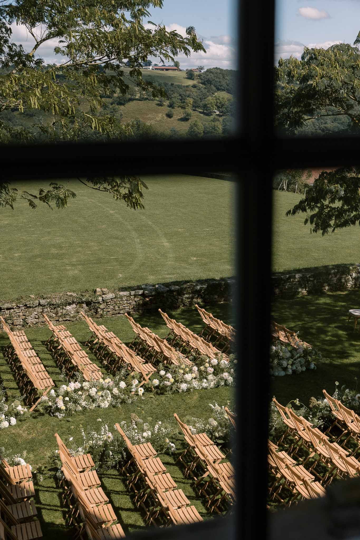 Outdoor ceremony setup viewed through window showing curved chair rows on lawn with white floral aisle borders