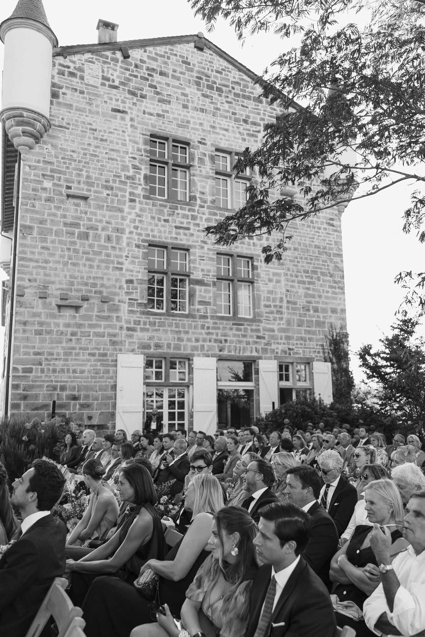 Black and white wide shot of outdoor wedding ceremony with guests seated before stone chateau with tower