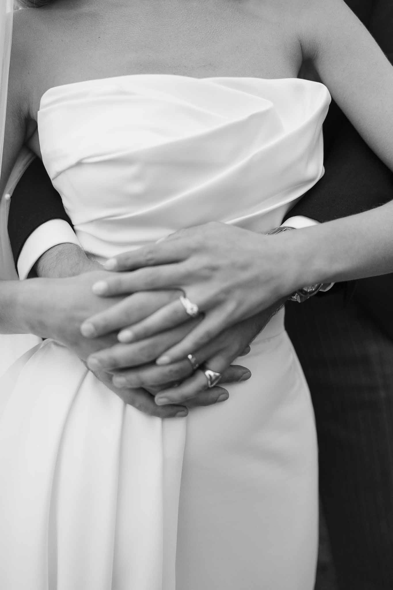 Black-and-white close-up of groom's hands around bride's waist showing engagement ring and satin gown