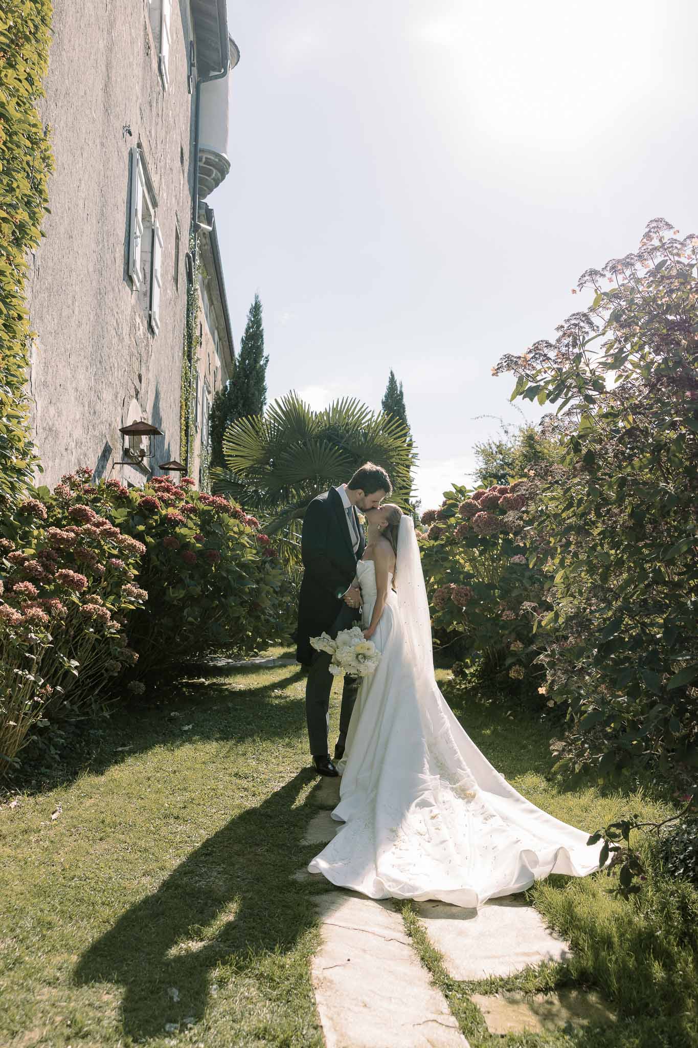 Couple kissing on stone path with cathedral train spread, bride holding white peony bouquet before ivy-covered manor